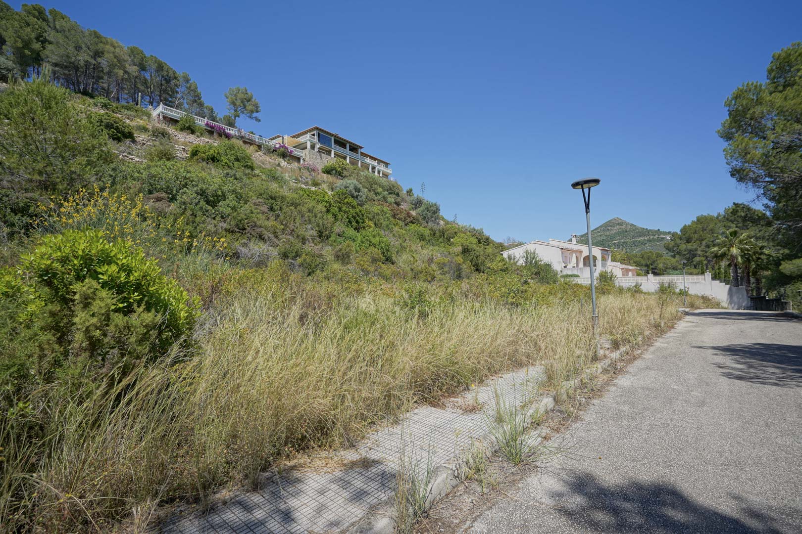 Straatzicht van de verhoogde woonwijk in Alcalalí, met geasfalteerde toegang, straatverlichting en de natuurlijke mediterrane heuvelomgeving.