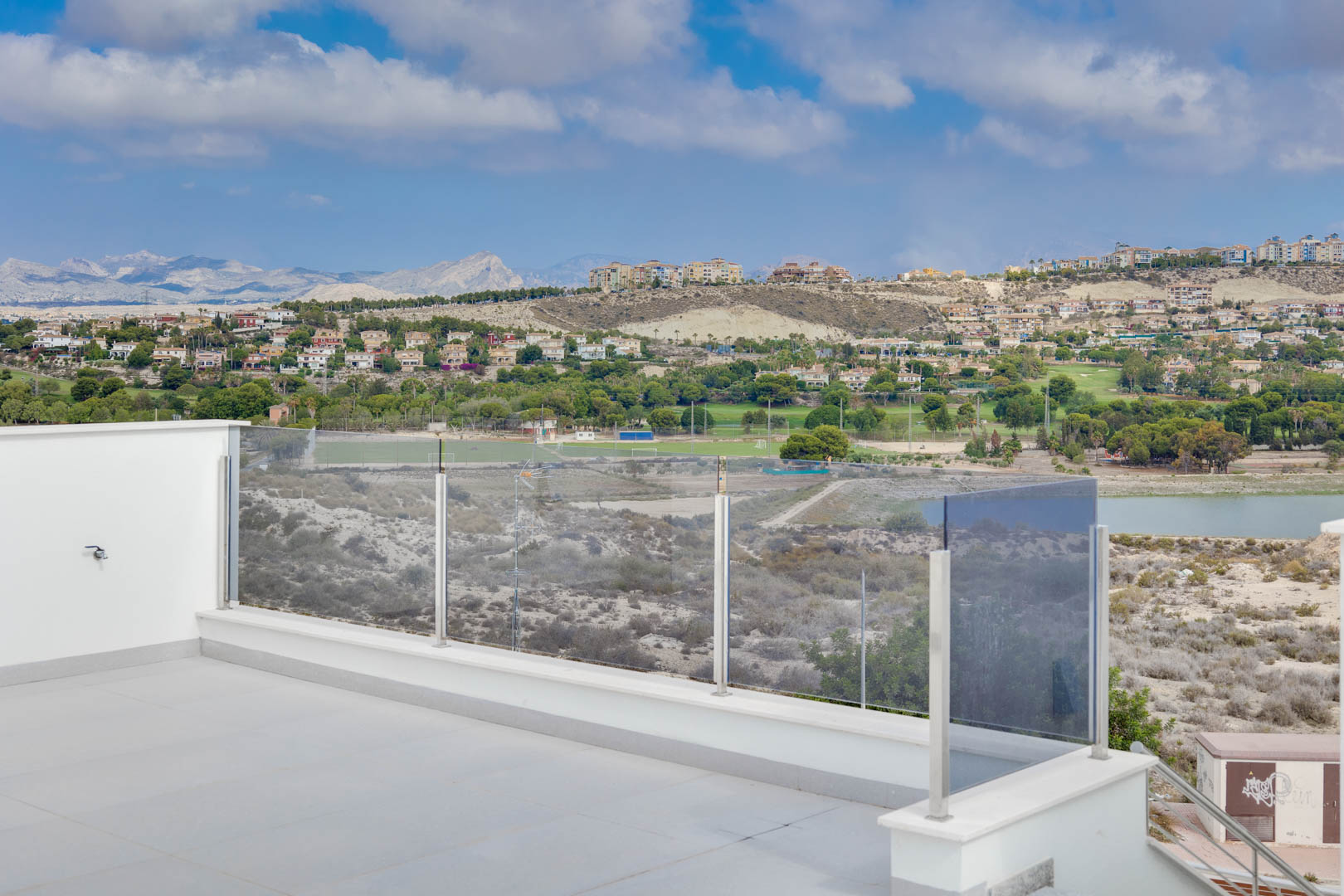 Bovenste terras met grote grijze vloertegels en glazen balustrades, met panoramisch uitzicht op de bergen, het meer en de omliggende golfbanen.