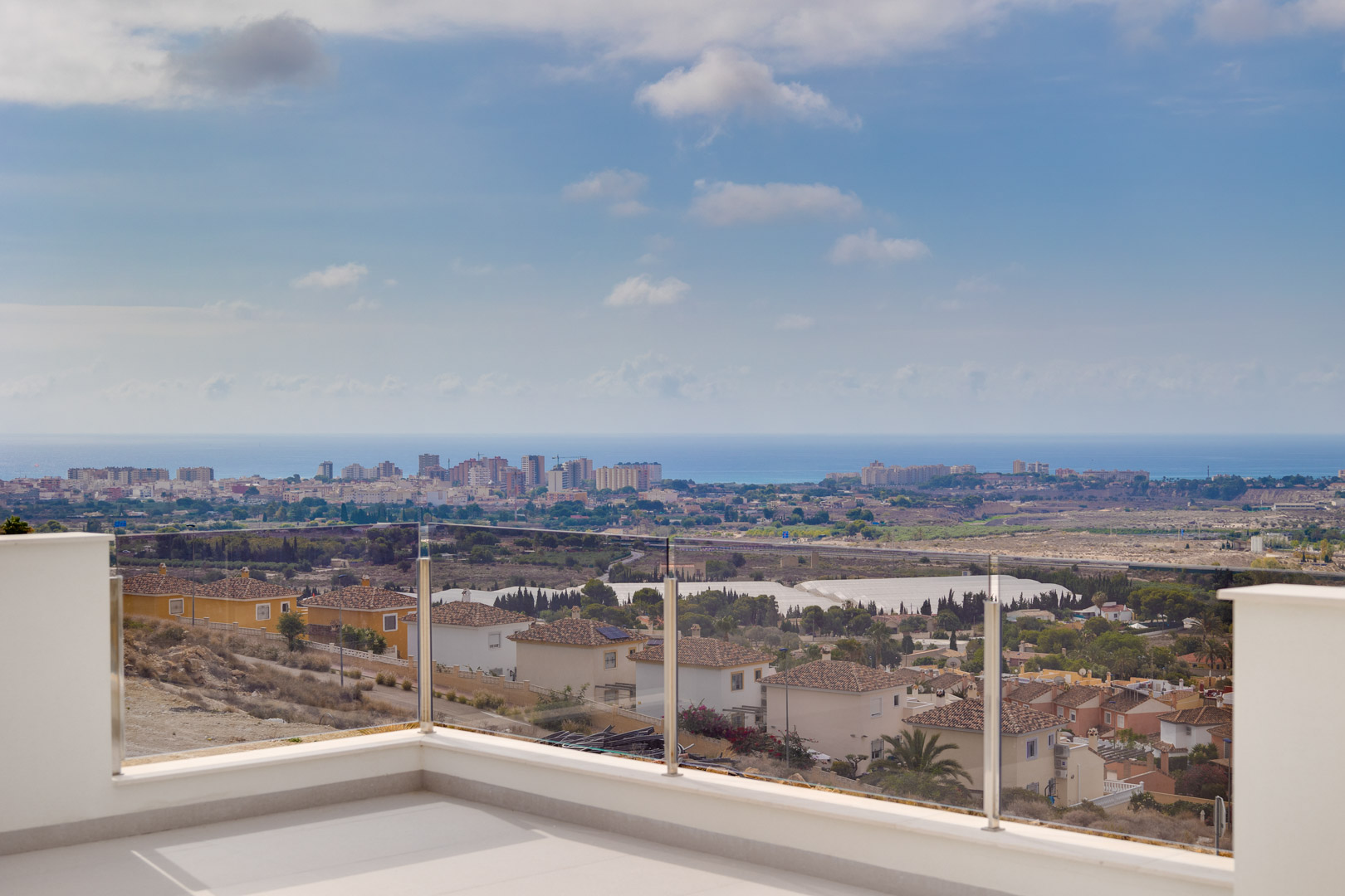 Bovenste terras met lichte keramische tegels en een glazen balustrade, met een onbelemmerd panoramisch uitzicht over de kustlijn en de Middellandse Zee.