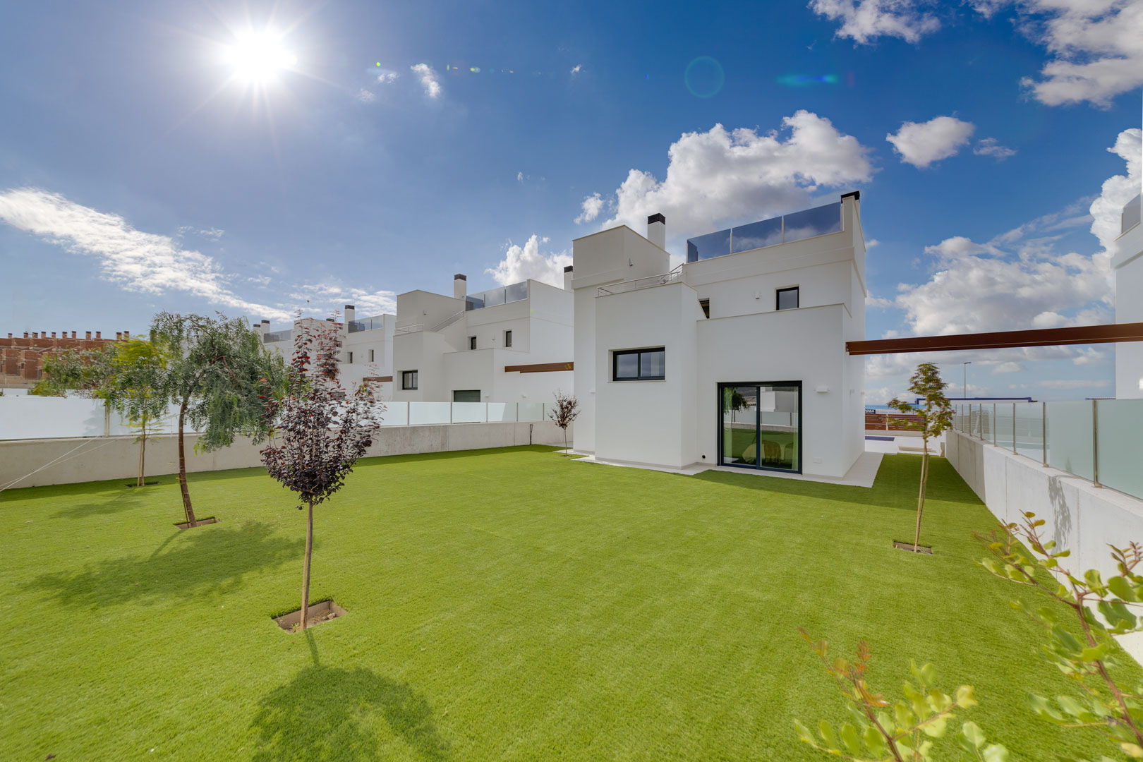 Buitenaanzicht van de villa met een minimalistische witte gevel, een ruim gazon met jonge bomen en glazen balustrades op het dak.