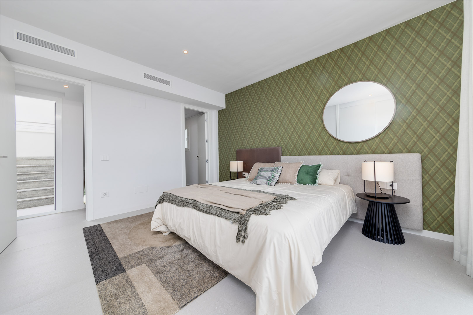 Eigentijdse slaapkamer met een olijfgroene accentmuur met patroon, ronde spiegel en geïntegreerde airconditioningroosters boven de neutrale vloer.