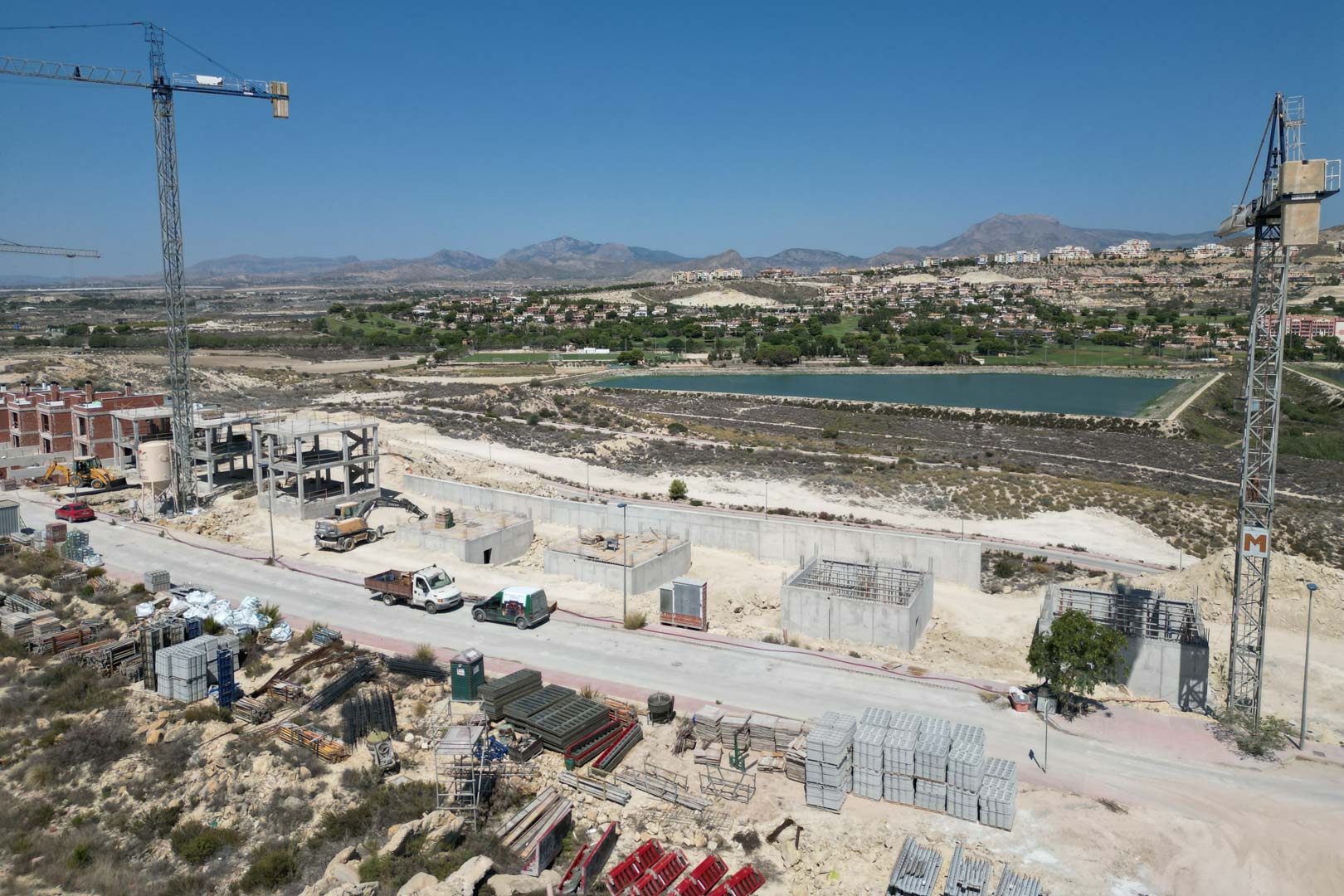 Luchtfoto van de villa's in aanbouw, met betonstructuren, kranen en een panoramisch uitzicht op het nabijgelegen meer en de bergen.