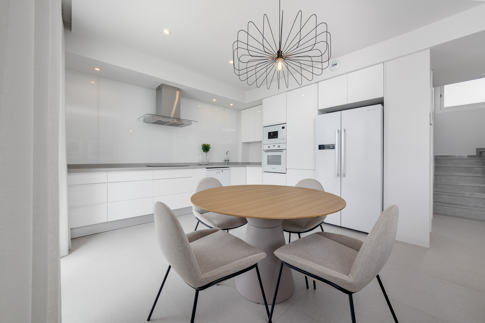 Minimalistische witte keuken met inbouwapparatuur, grijs werkblad en eethoek met een ronde houten tafel en moderne draadlamp.