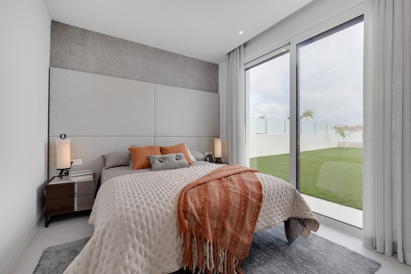 Moderne slaapkamer met gestoffeerd hoofdbord, aardetinten en grote glazen schuifdeuren die directe toegang bieden tot de privétuin.