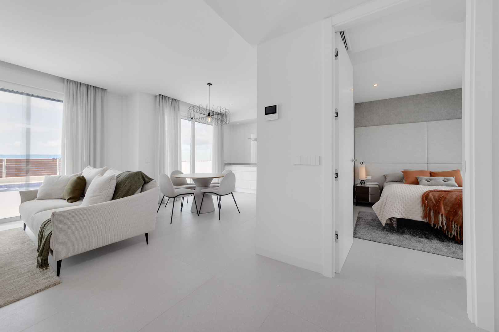 Open woon- en eetkamer met grote vloertegels, modern meubilair en doorkijk naar een slaapkamer met aardetinten en een gestoffeerd hoofdbord.