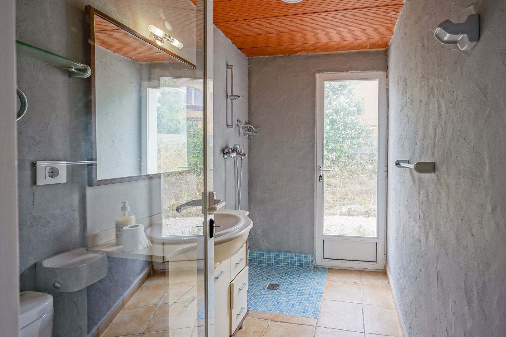 Badkamer met grijze pleisterwanden, oranje lamellenplafond en een douchegedeelte met blauwe mozaïektegels. Voorzien van een glazen deur naar de tuin.