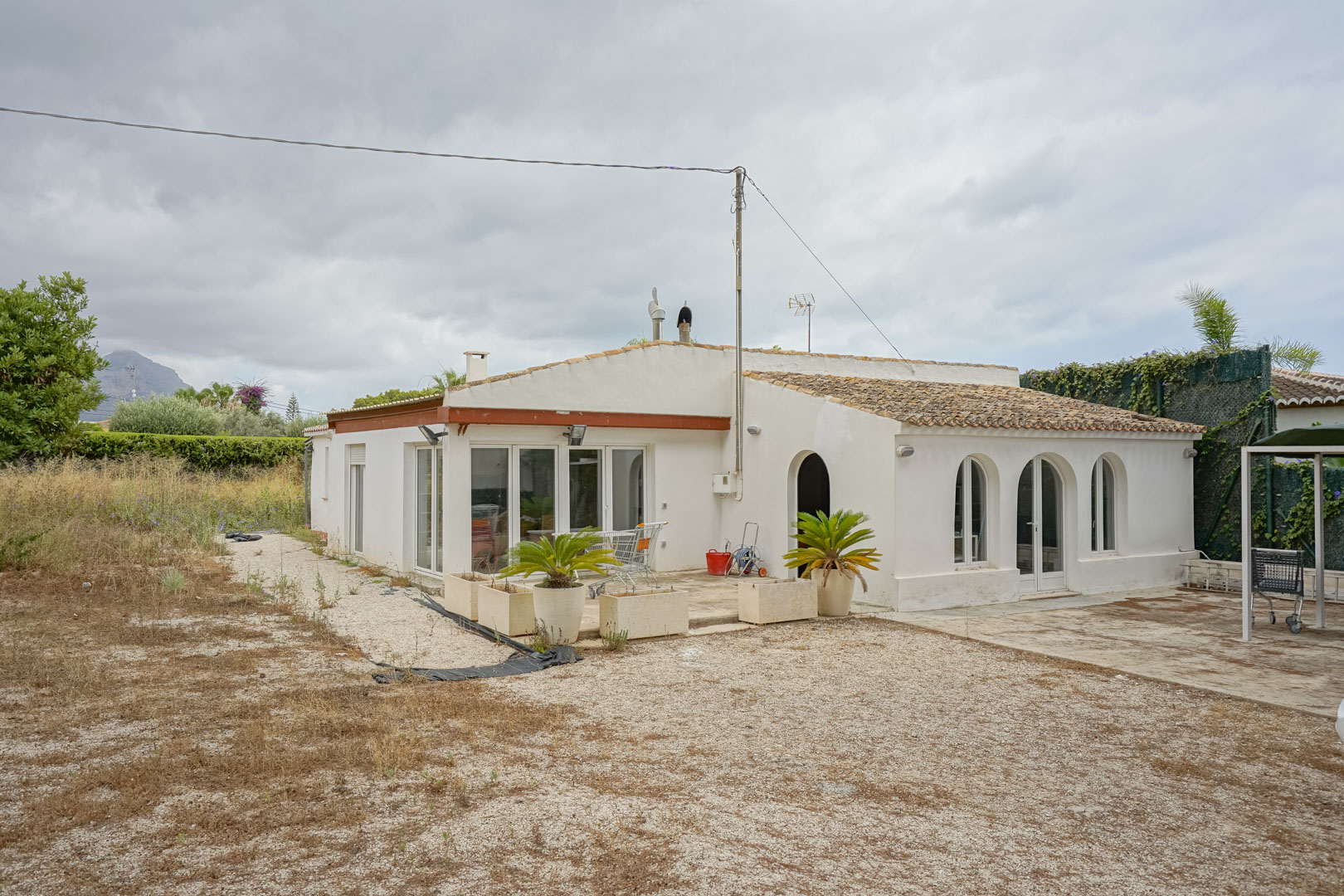 Buitenaanzicht van de mediterrane villa met traditionele bogen, wit schrijnwerk en een pannendak op een uitgestrekt vlak perceel met uitzicht op de bergen.