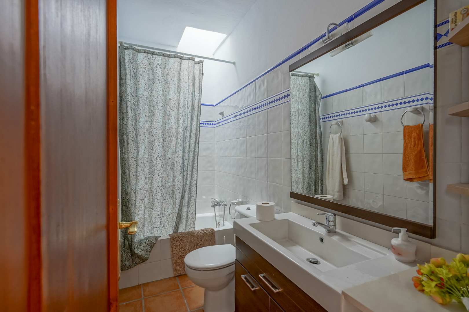 Functionele badkamer met wit-blauwe betegeling, een donkerhouten wastafelmeubel en een ligbad voorzien van een dakraam voor natuurlijk lichtinval.