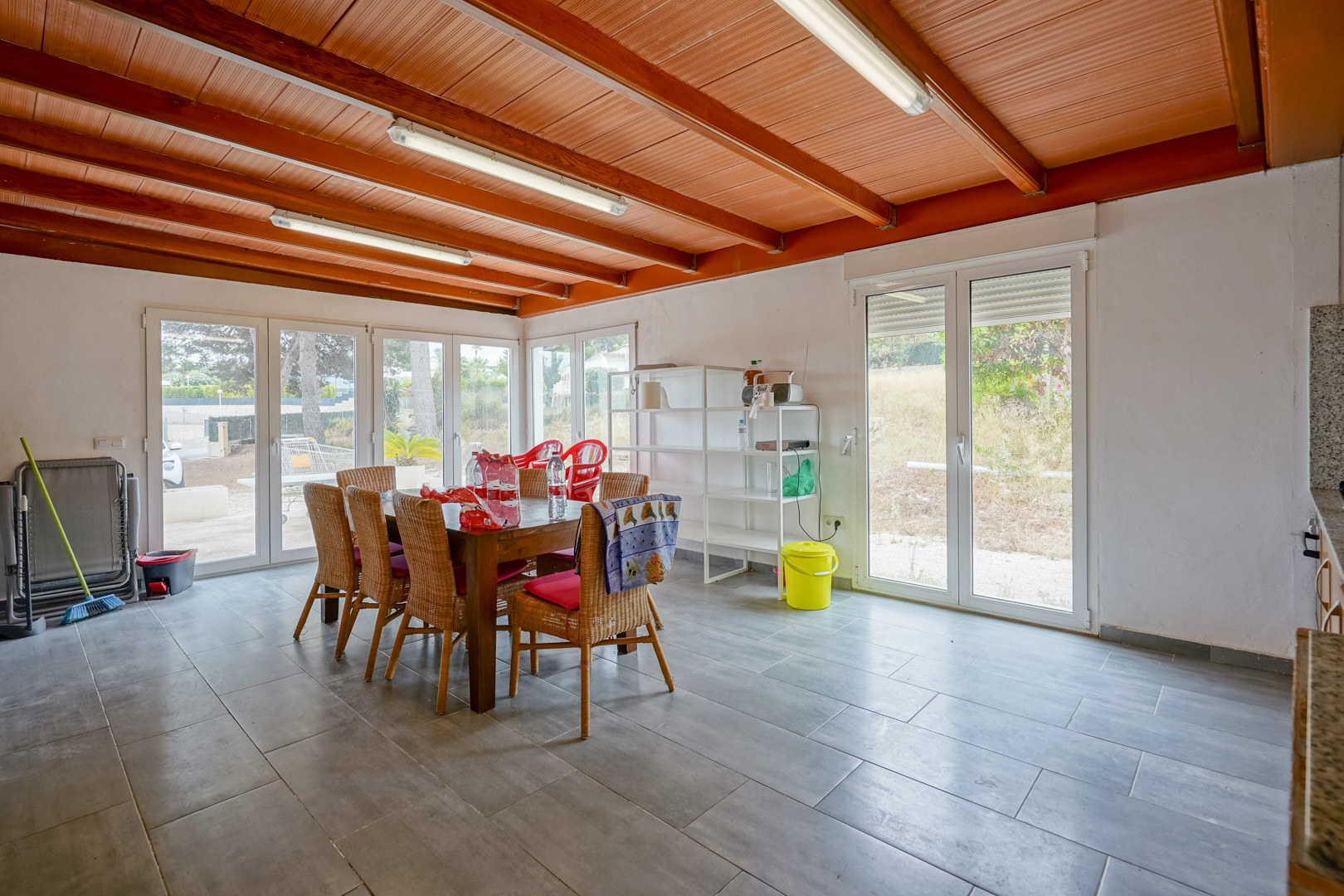 Lichte eetkamer met een plafond van houten balken, grijze tegelvloer en grote witgekleurde glazen deuren die directe toegang bieden tot de tuin.