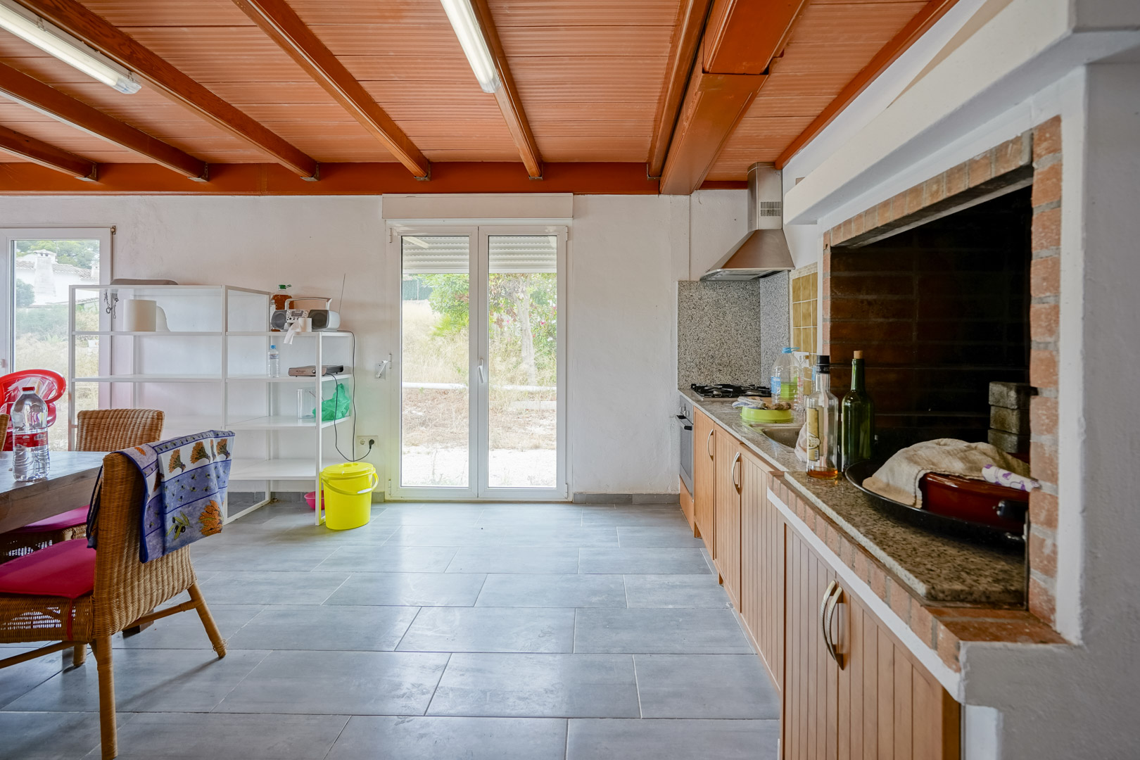Rustieke keuken met houten balkenplafond, granieten werkblad en een ingebouwde stenen barbecue, met directe toegang tot de tuin via dubbele glazen deuren.