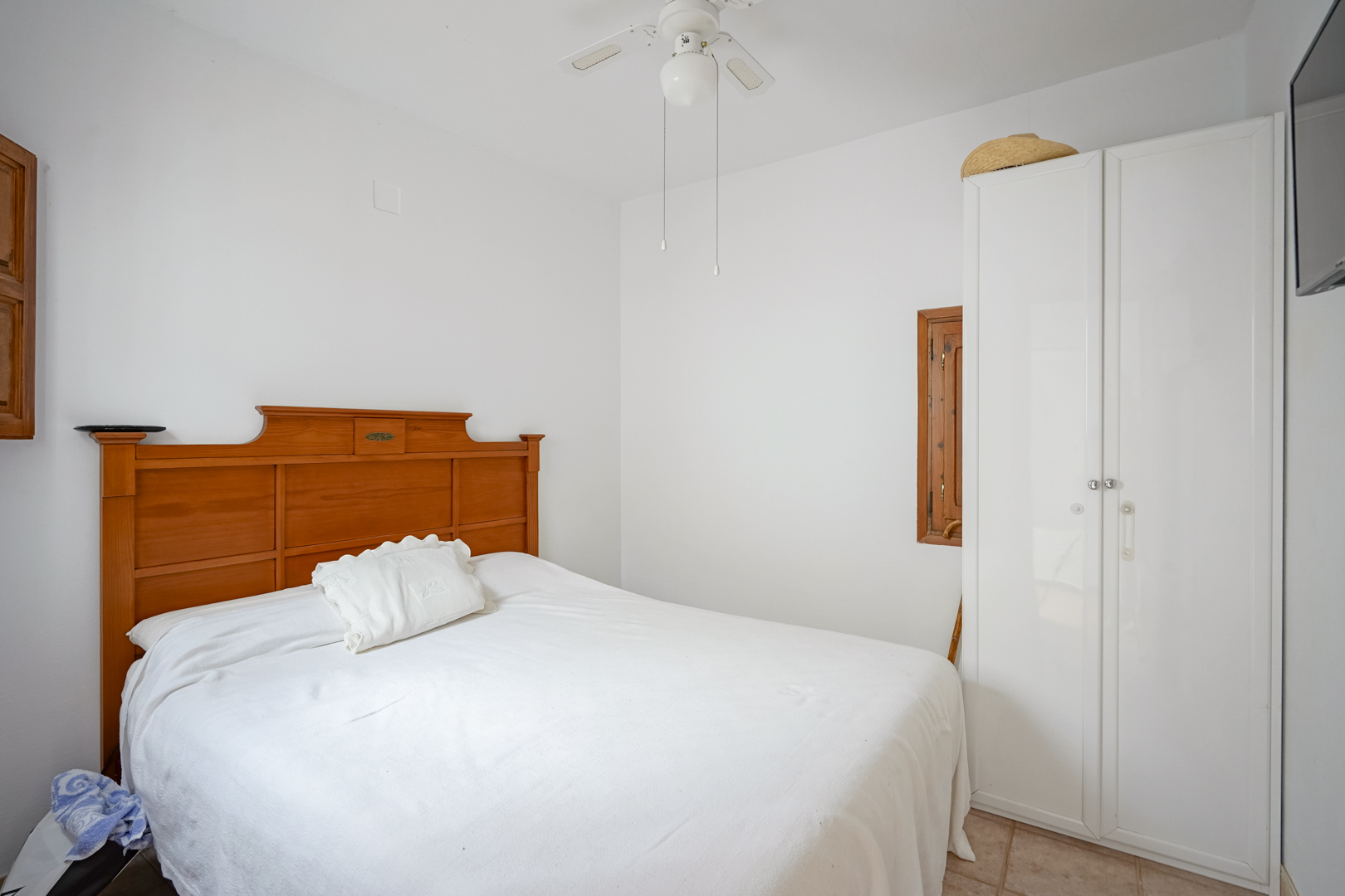 Slaapkamer met tweepersoonsbed, houten hoofdbord, witte kast en plafondventilator. Witte muren en een tegelvloer zorgen voor een lichte uitstraling.