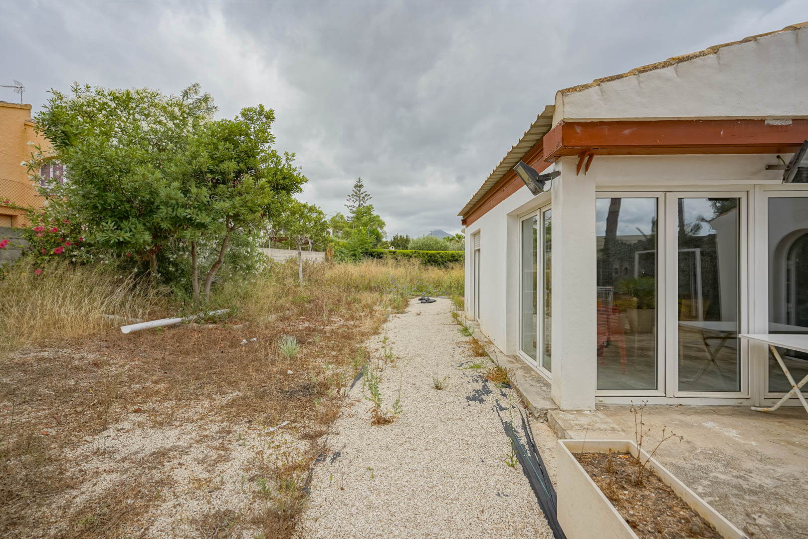 Zijgevel van de villa met grote witte kunststof raampartijen en toegang tot een uitgestrekt natuurlijk perceel met mediterrane begroeiing.