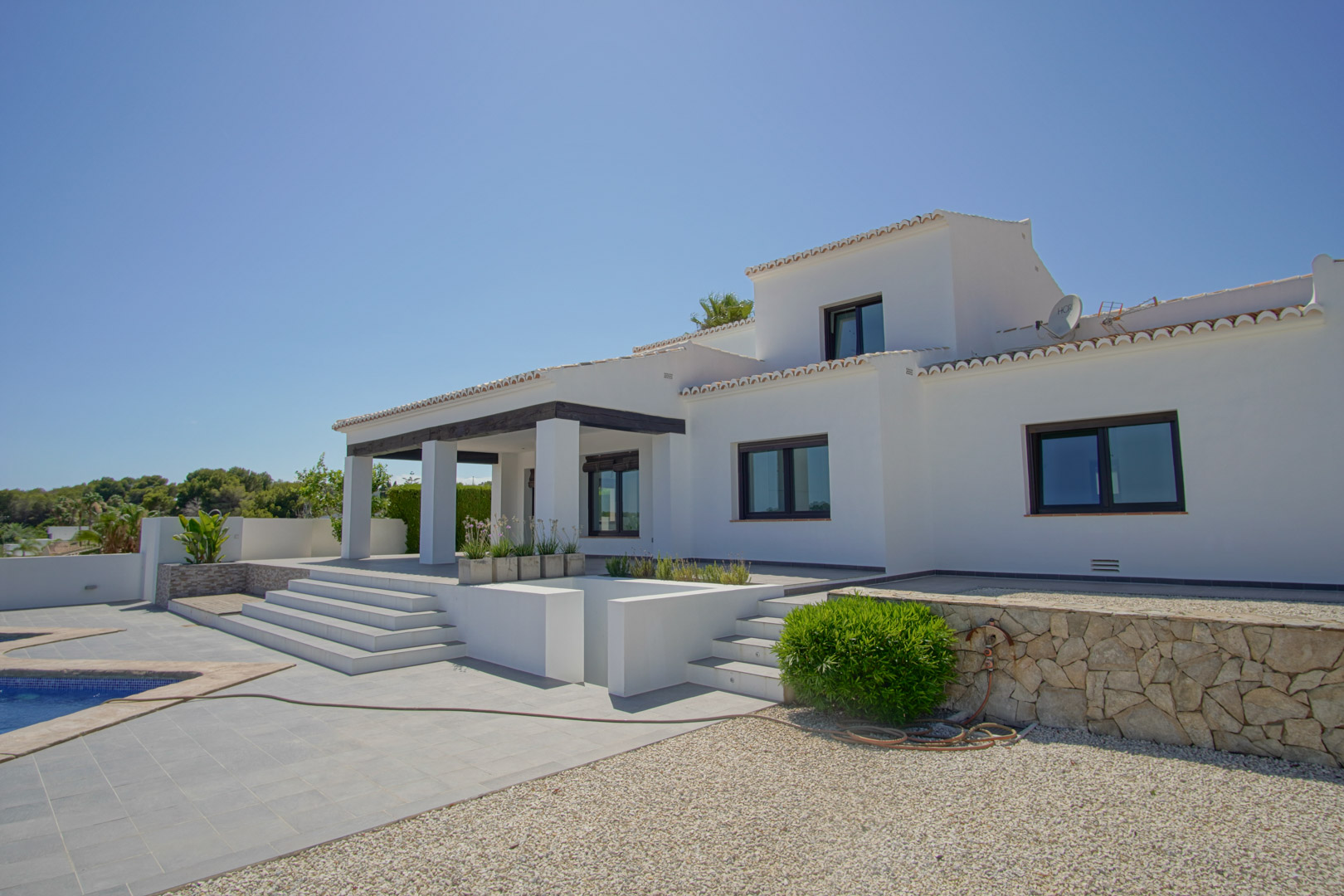 Buitenaanzicht van de mediterrane villa met witte gevel, veranda met donkere balken en een gelaagd terras dat aansluit op het zwembad en de stenen muren.