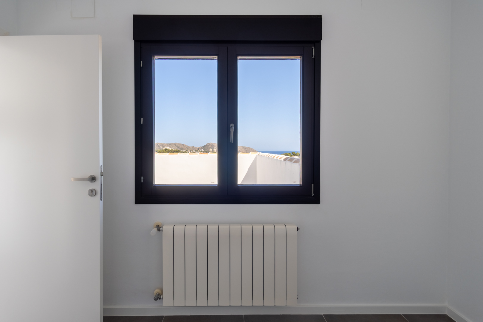 Donker aluminium raam met dubbele beglazing en uitzicht op de zee en Cabo de Moraira, geplaatst boven een witte radiator in een lichte, strak afgewerkte kamer.