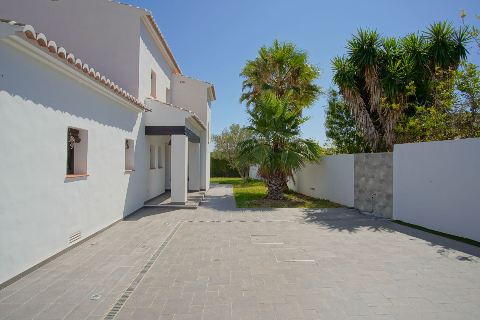 Geplaveide oprit met grijze tegels, grondspots en een mediterrane tuin met palmbomen grenzend aan de witte gevel van de woning.