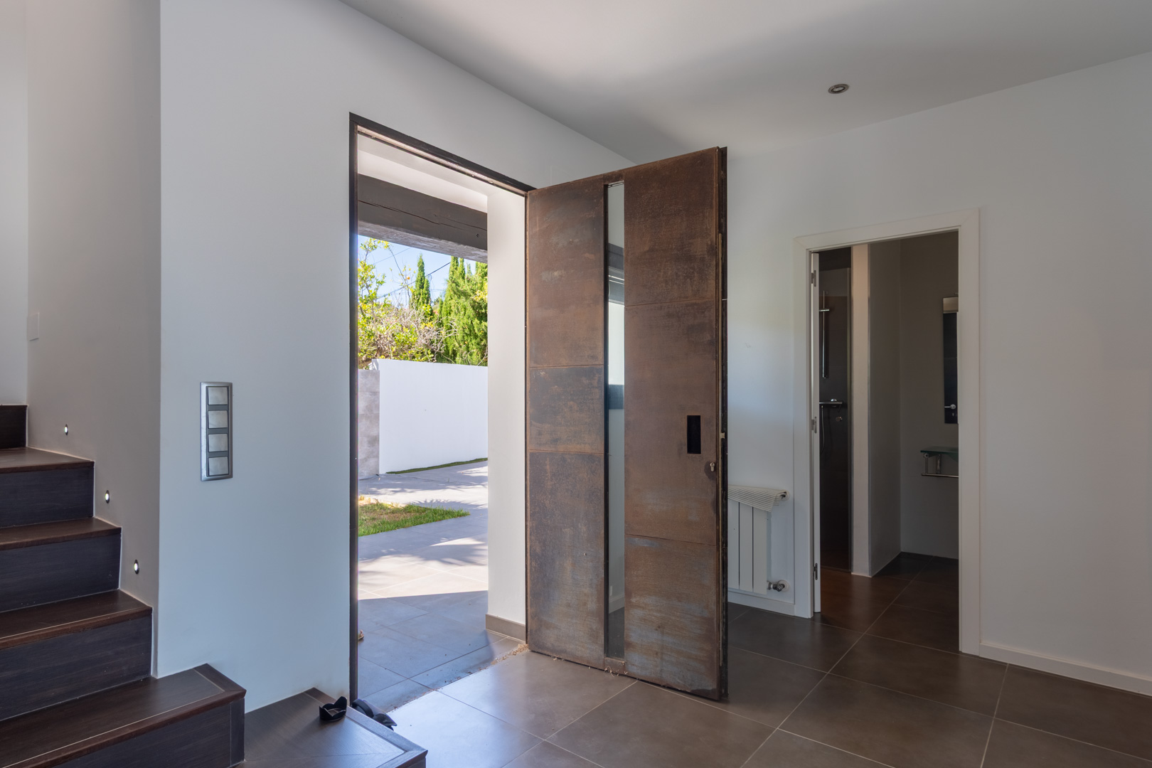Hoofdentree met een pivotdeur in cortenstaal-look en verticaal glas. De hal beschikt over donkere vloertegels, een trap met LED-verlichting en toegang tot een badkamer.