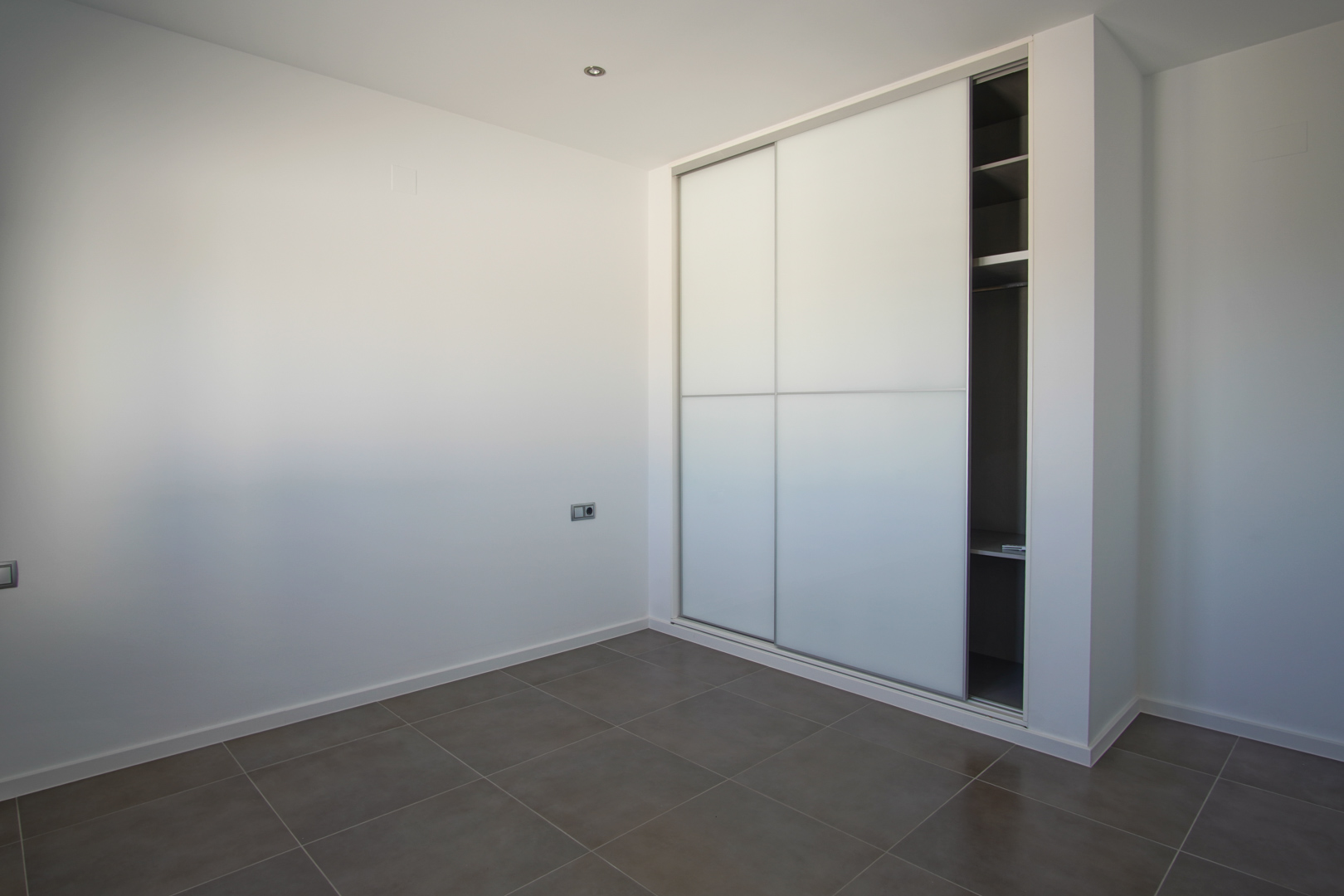 Inbouwkast met witte schuifdeuren en interne planken, geplaatst in een moderne ruimte met grijze vloertegels en witte muren.