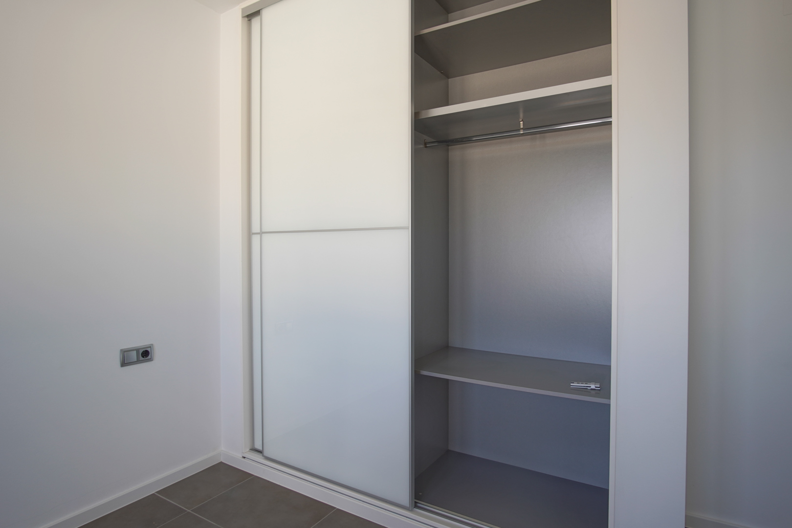Ingebouwde kledingkast met witte schuifdeuren en grijs interieur, voorzien van legplanken en een hangrail in een minimalistische kamer.