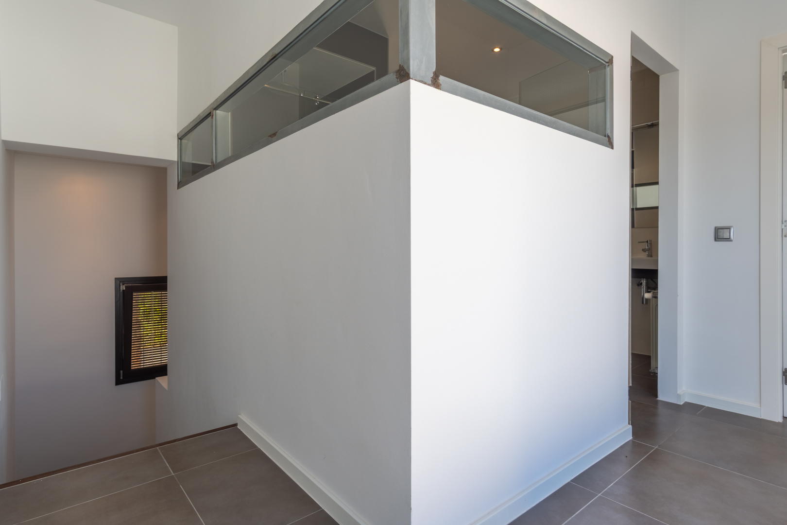 Interieurhal met grijze vloertegels, een scheidingswand met glazen bovenpanelen in metalen frame en toegang tot de trap en de badkamer.