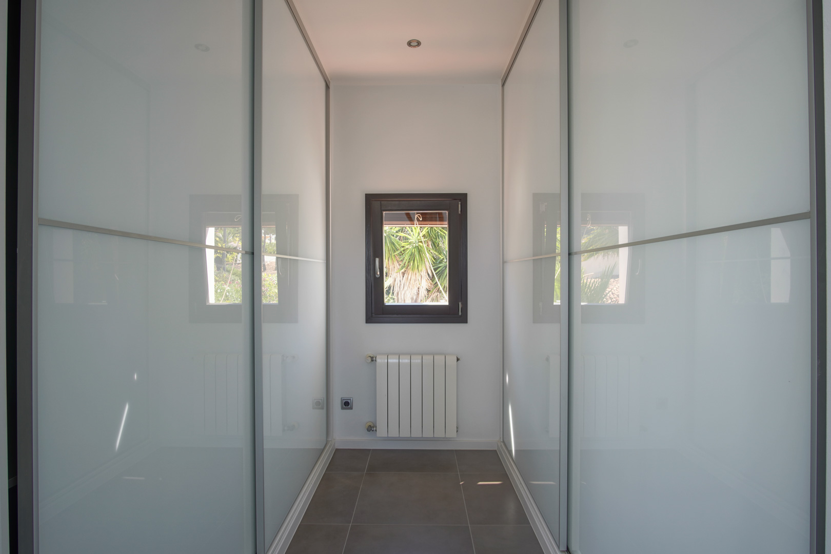 Kleedruimte met ingebouwde kasten met matglazen schuifdeuren, een radiator onder het raam en een strakke, donkere tegelvloer.