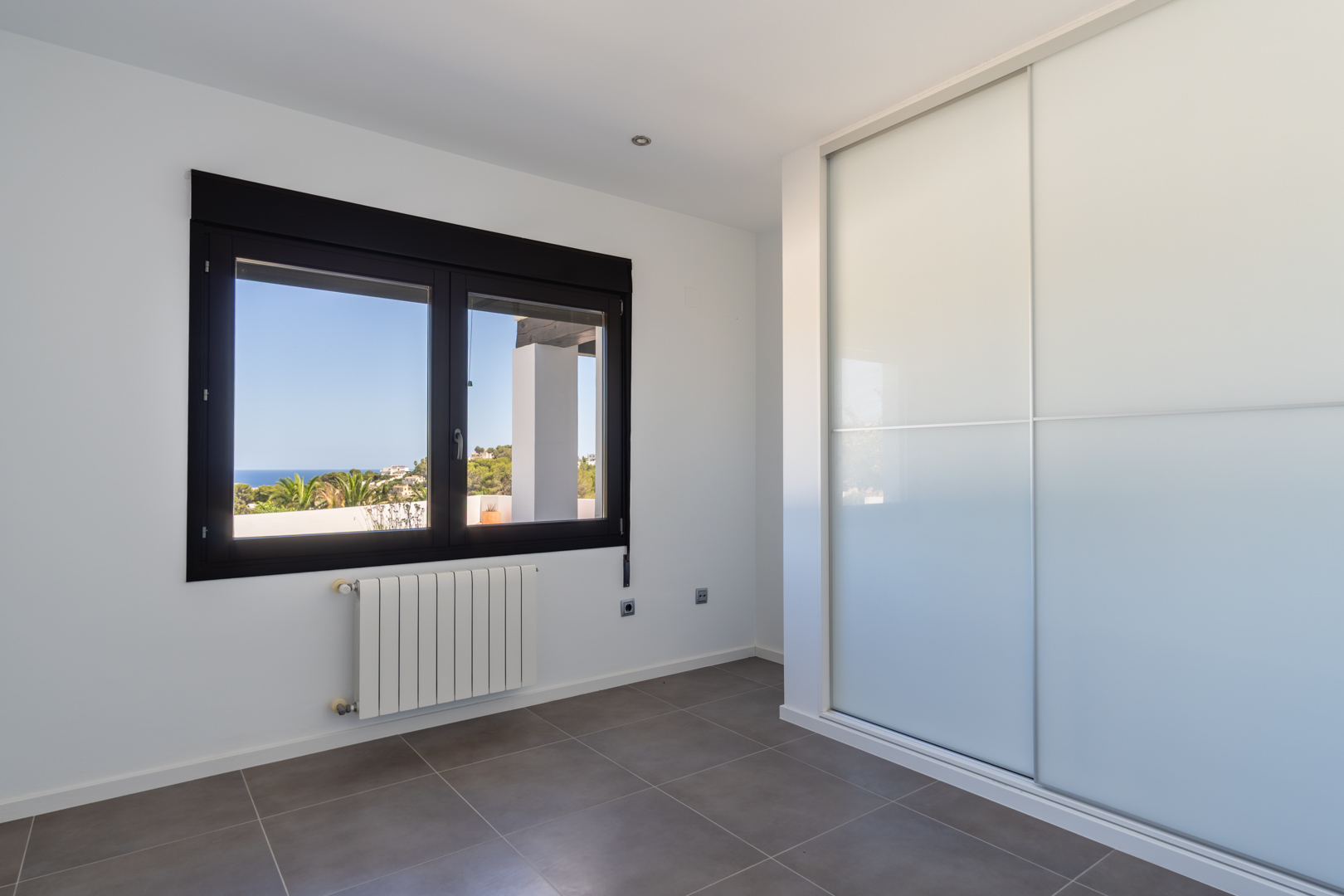 Lichte slaapkamer met inbouwkast met schuifdeuren, radiator en een raam met donker profiel dat uitzicht biedt op de Middellandse Zee en de kustlijn.