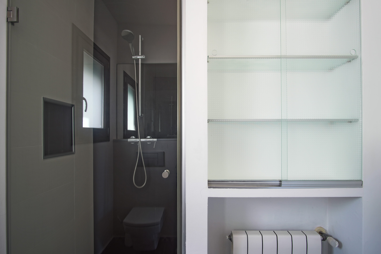 Moderne badkamer met donkergrijze tegels, inloopdouche met chromen kranen en een ingebouwde kast met glazen planken naast een radiator.