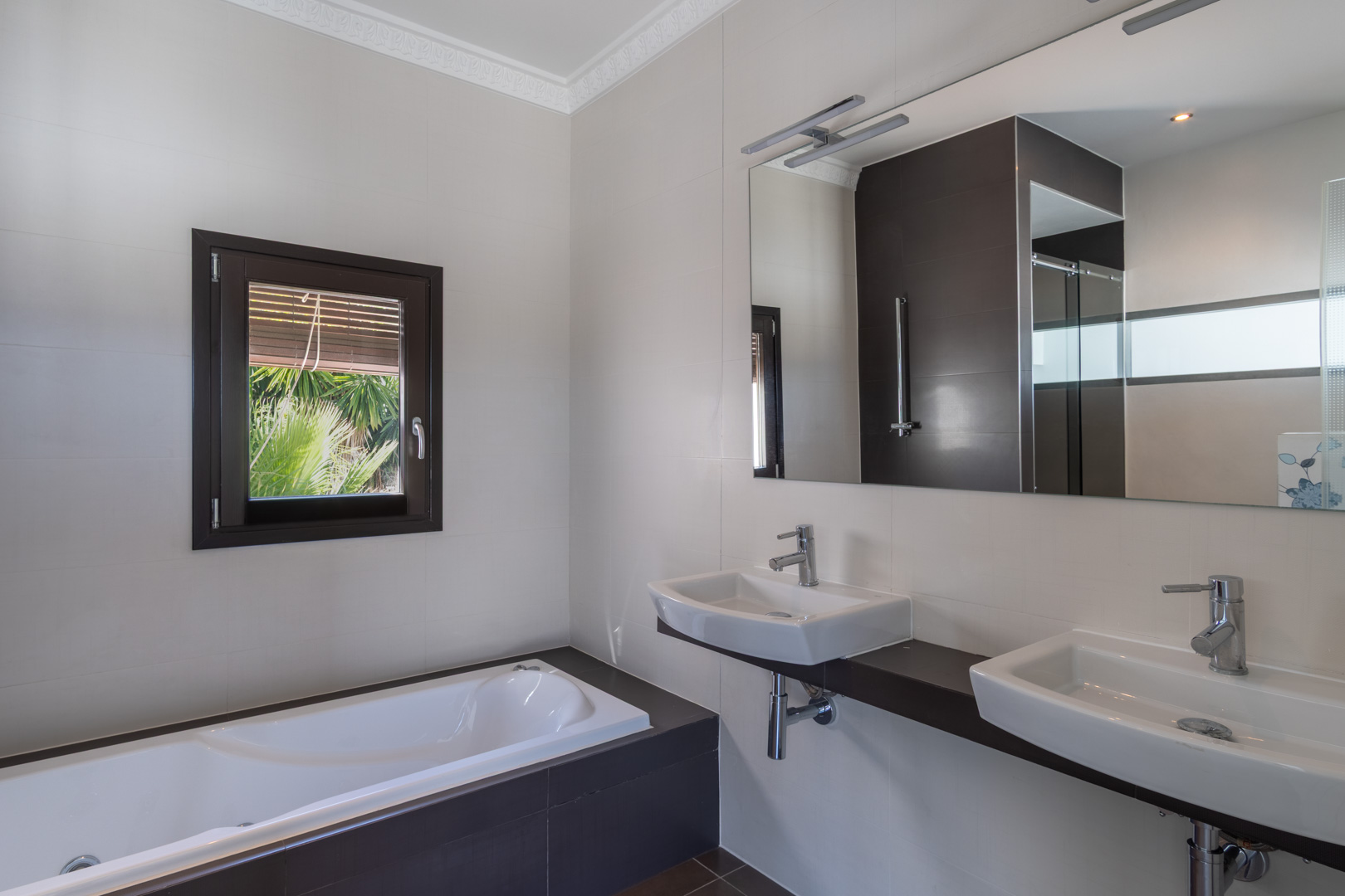 Moderne badkamer met dubbele wastafel op een donker blad, ingebouwd ligbad en raam met houtlook kozijn dat zorgt voor natuurlijk licht en uitzicht op de tuin.