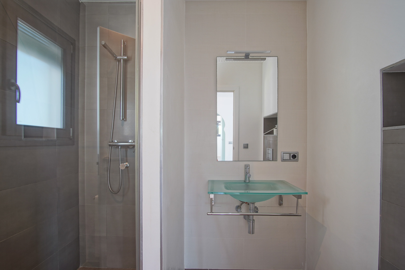 Moderne badkamer met een doorschijnende glazen wastafel, spiegel met verlichting en een inloopdouche met donkergrijze tegels en een glazen wand.