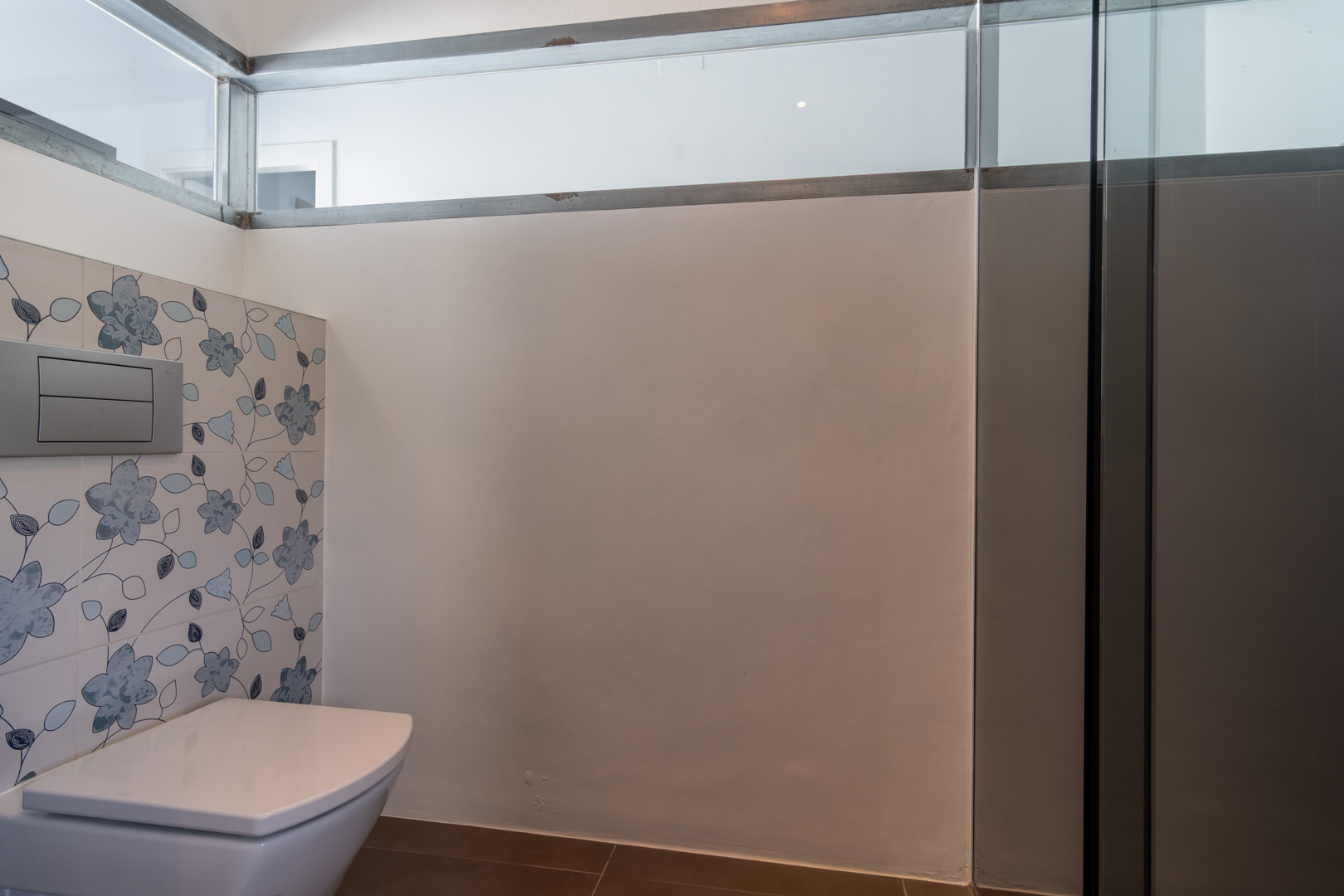 Moderne badkamer met hangend toilet, decoratieve tegels met blauw bloemenmotief en een glazen bovenpaneel voor natuurlijke lichtinval.
