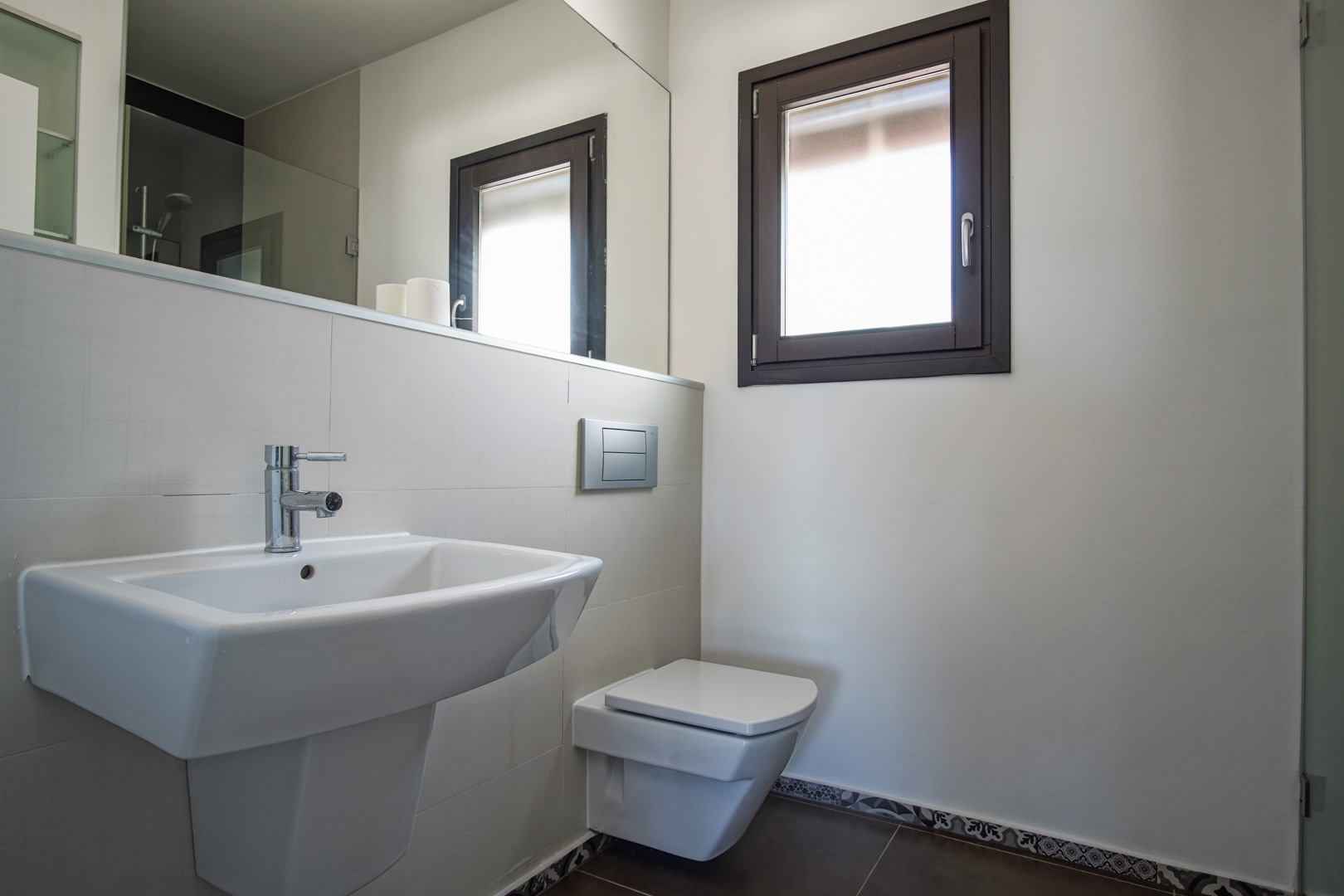 Moderne badkamer met hangend toilet, witte keramische wastafel en aluminium raam. Voorzien van een grote spiegel en donkere vloertegels met decoratieve rand in hydraulische stijl.