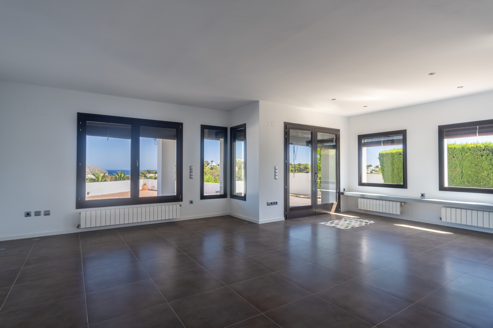 Ruime woonkamer met donkere tegelvloer, radiatoren en aluminium kozijnen met houteffect die uitkijken over de zee en de kustlijn van Moraira.