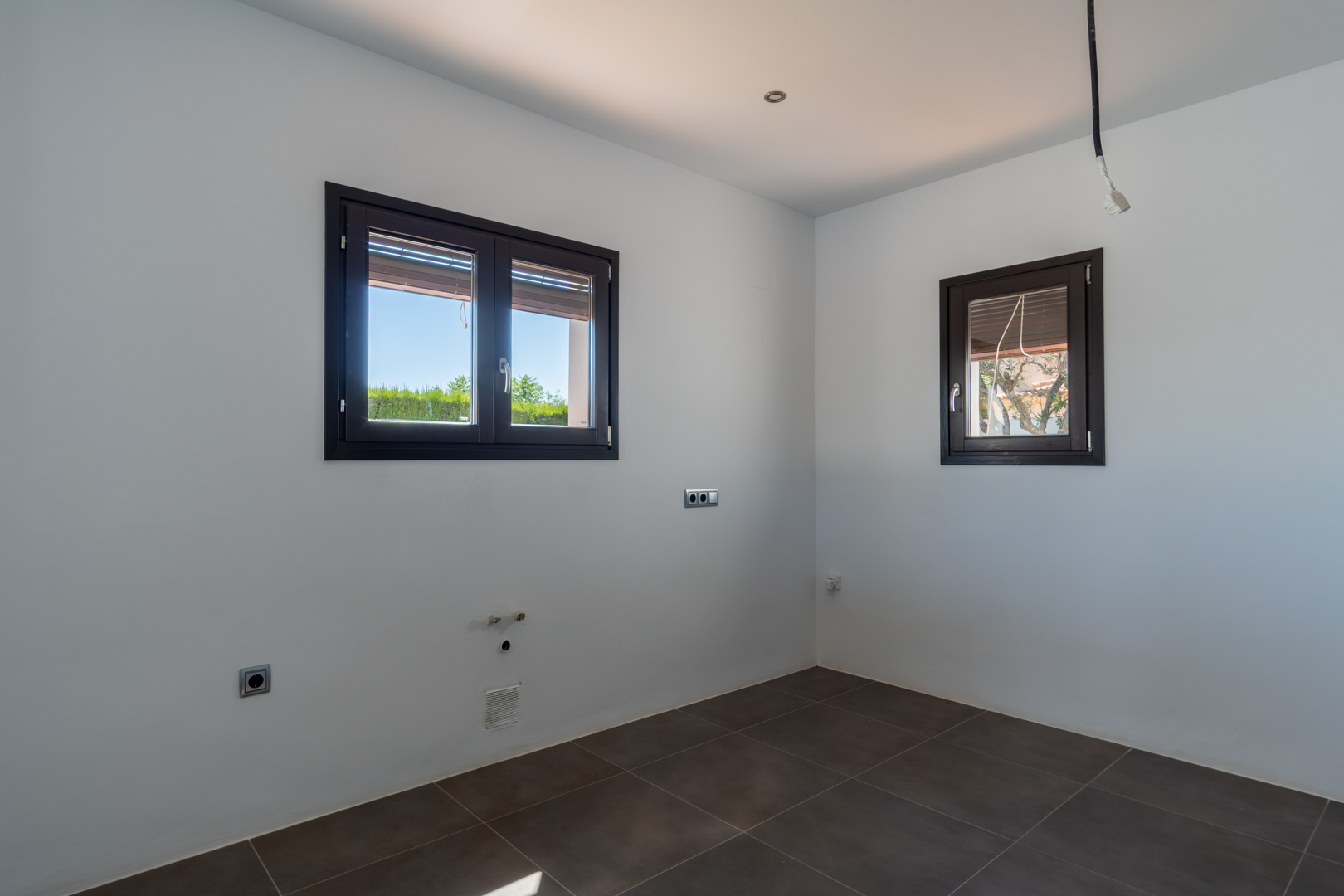 Ruimte voorbereid voor keukeninstallatie met donkere vloertegels, aluminium ramen met houtlook en aanwezige aansluitingen voor water en elektriciteit.