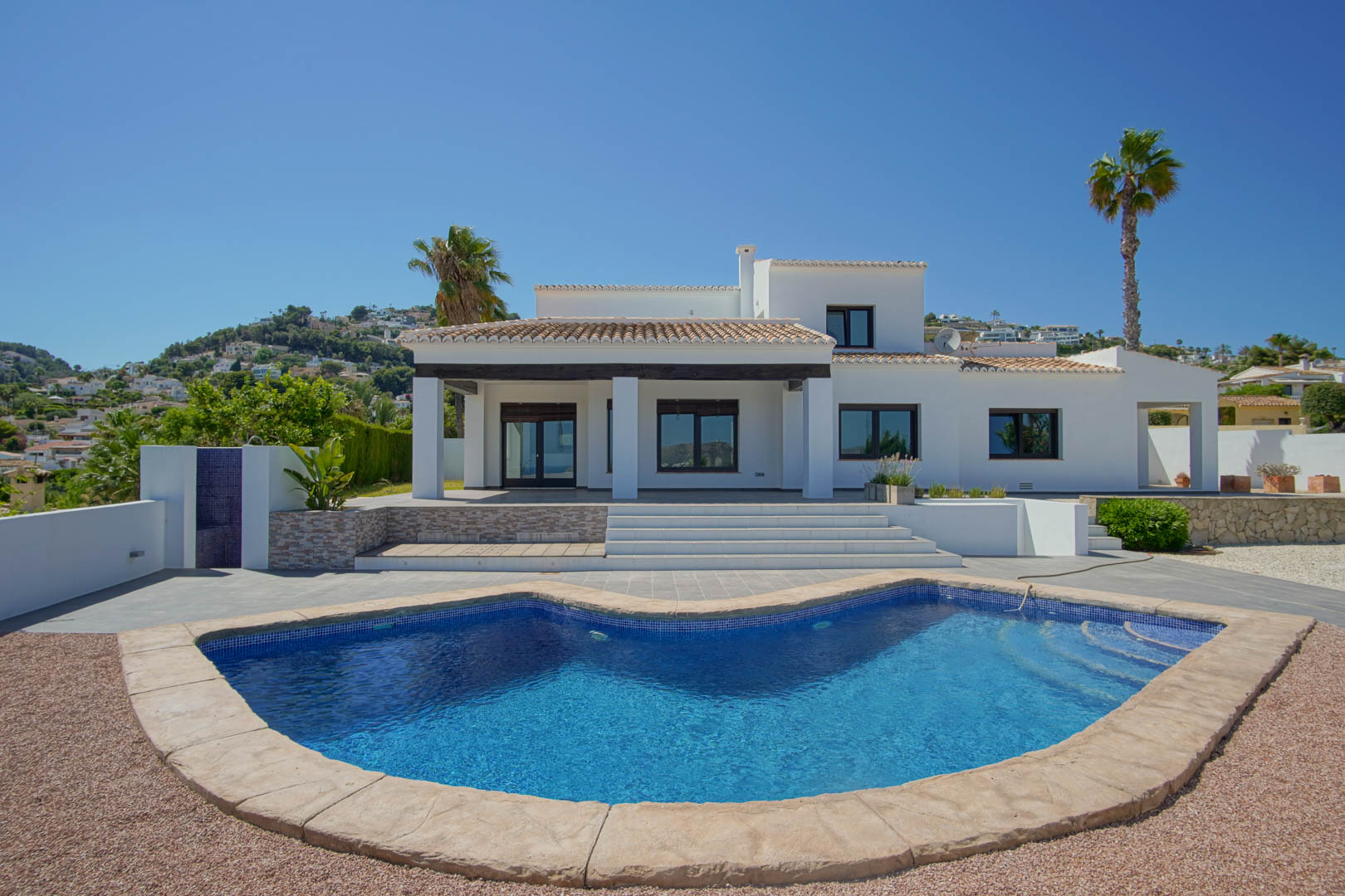 Voorgevel van de witte mediterrane villa met een niervormig zwembad, overdekt terras met donkere houten balken en ruime zonneterrassen.