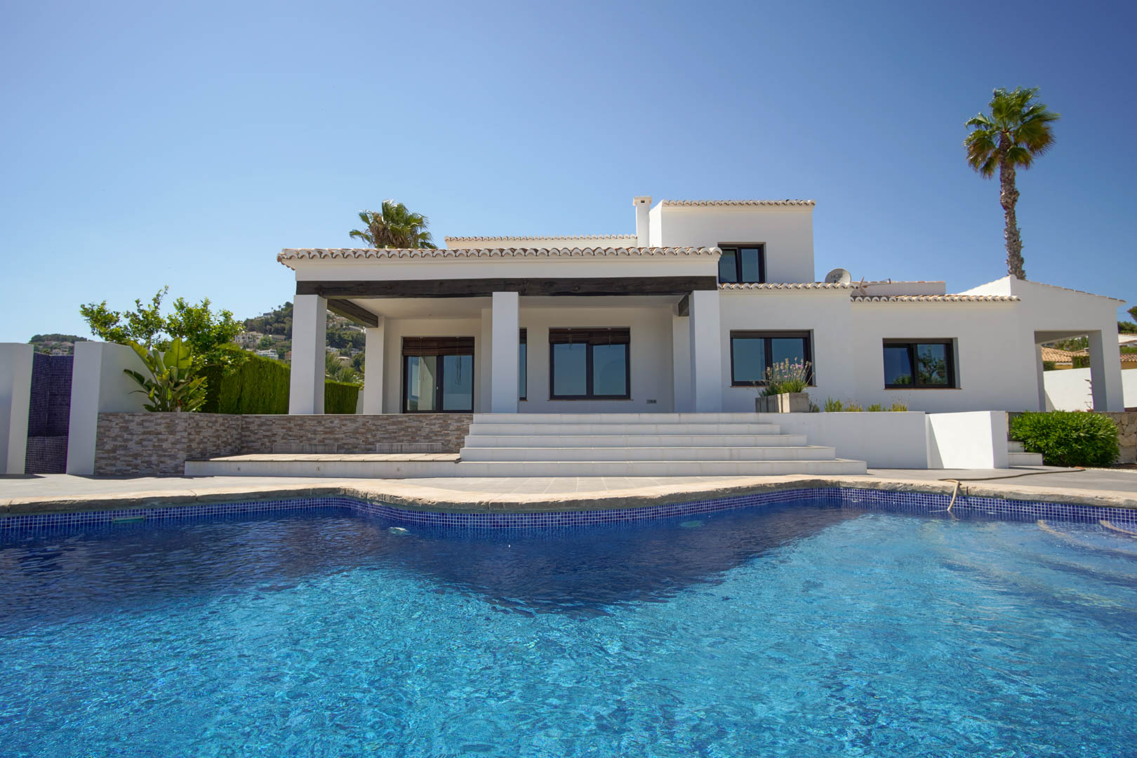 Witte mediterrane villa met een blauw betegeld zwembad. Het exterieur biedt een overdekt terras met donkere houten balken en brede trappen naar de woning.