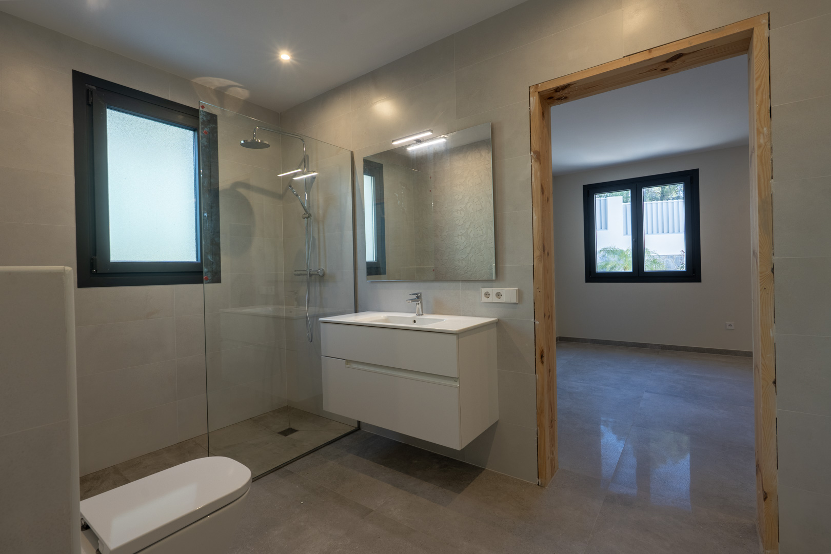 Detail van een moderne badkamer: douche met glazen wand, zwevend wastafelmeubel en raam met diffuus licht.