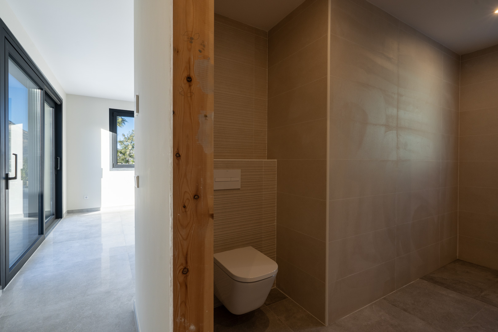 Detail van toiletruimte met zandkleurige tegels en een onafgewerkte houten deurpost, uitkijkend op de hal.