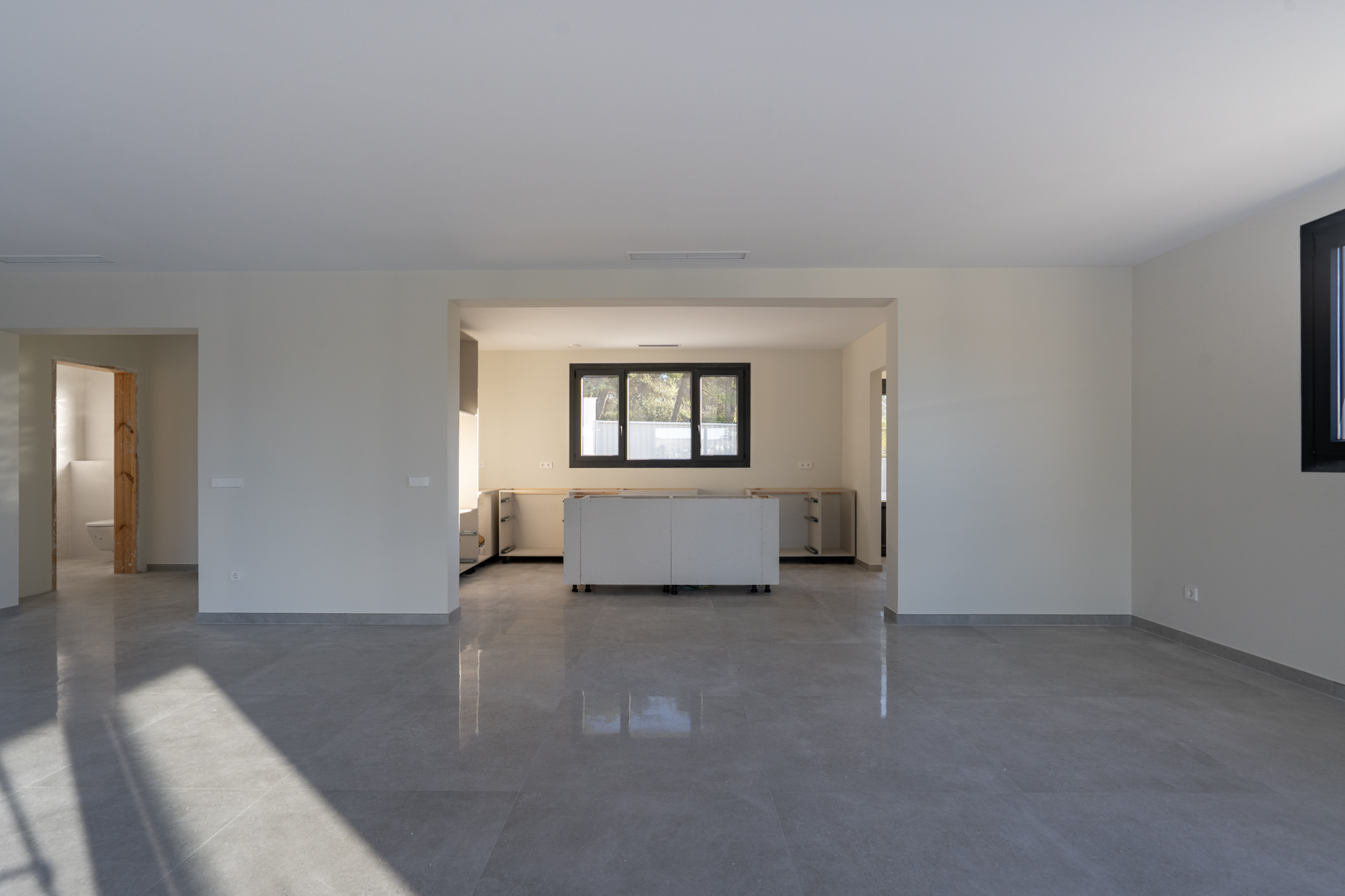 Open interieur met grijze tegelvloer. Keuken in aanbouw met witte onderkasten en raam met zwarte kozijnen.