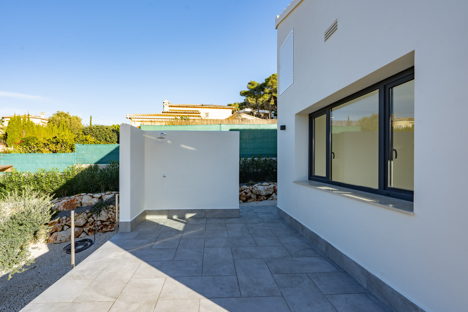 Patio met buitendouche en grijze tegels, naast een witte muur met dubbel glas.