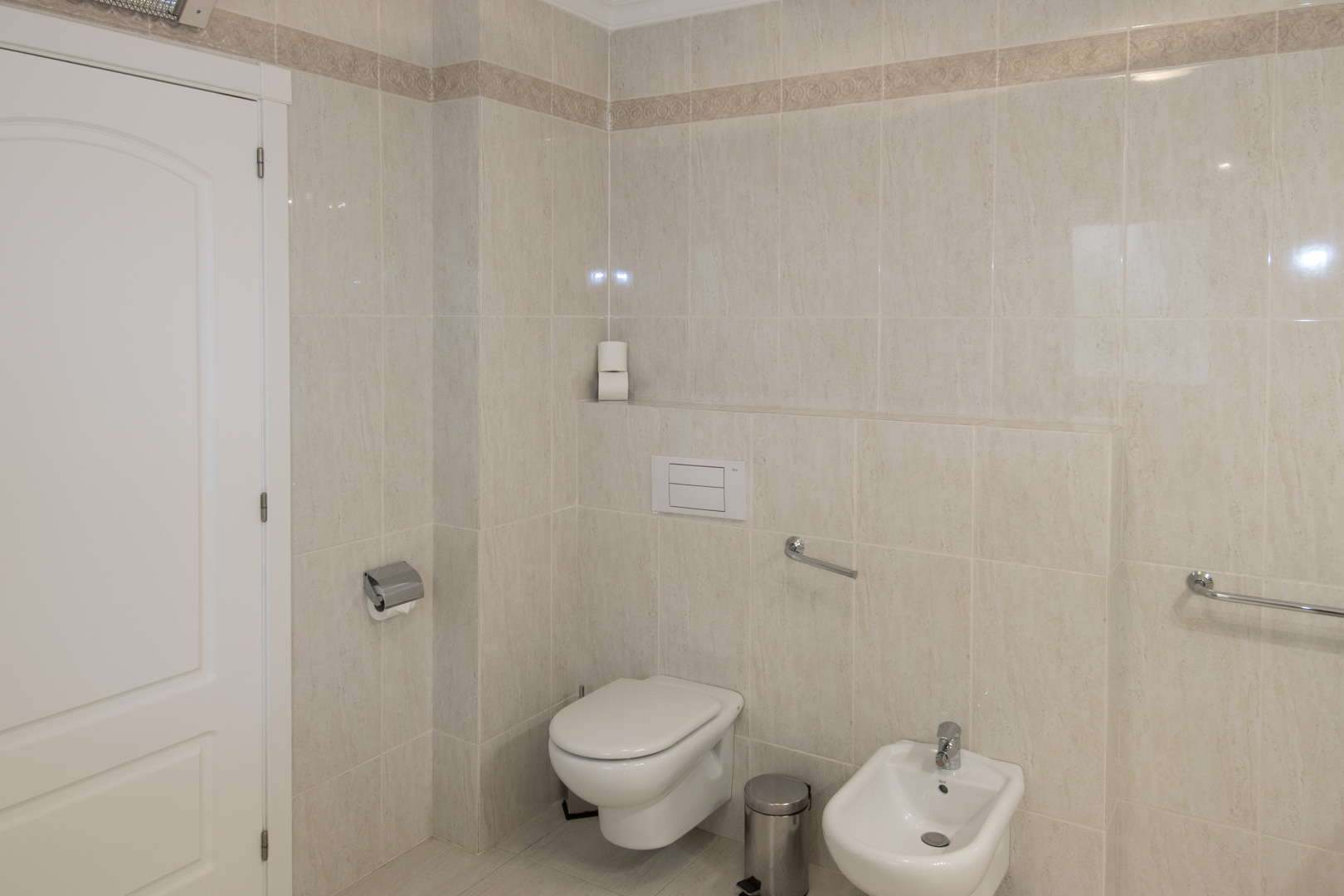 Badkamer met crèmevloer- en wandtegels, voorzien van een hangend toilet, bidet en chromen veiligheidsbeugels aan de wanden.