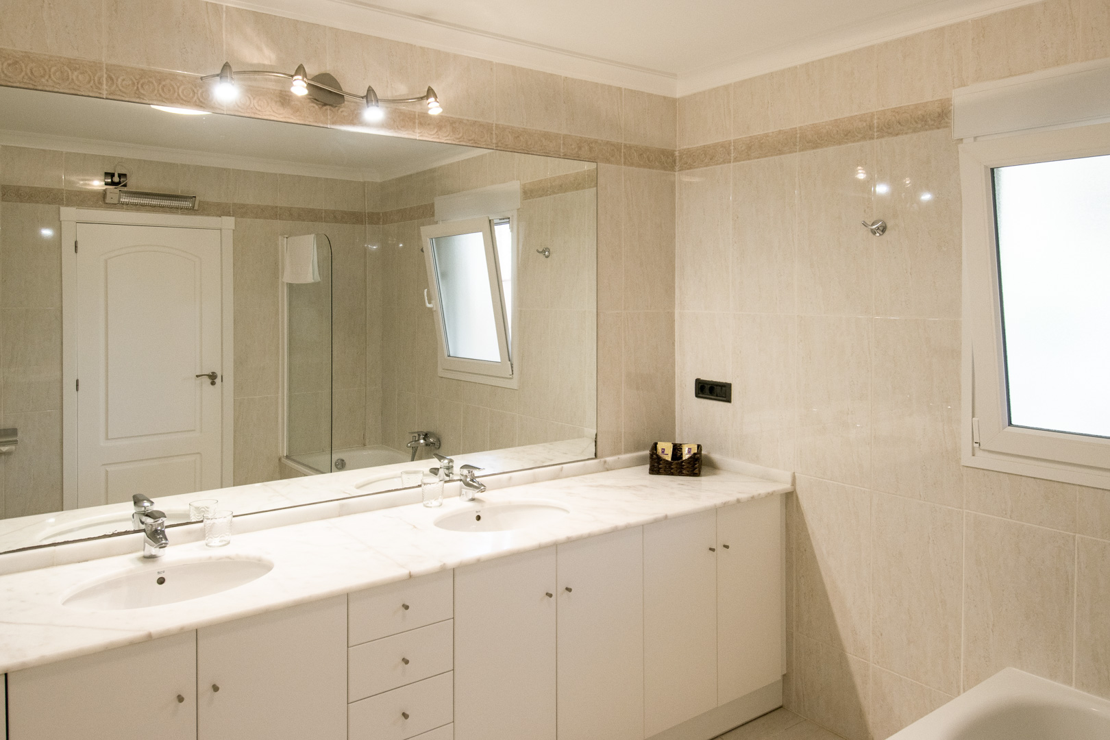 Badkamer met een dubbele wastafel op een wit marmeren blad en een kamerbrede spiegel. De wanden zijn afgewerkt met beige tegels en een subtiele decoratieve rand.
