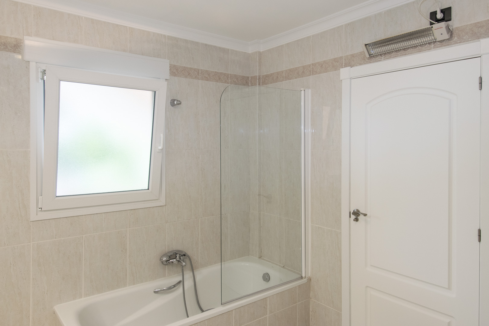 Badkamer met ligbad en glazen wand, betegelde muren met een decoratieve bies, een PVC-raam met matglas en een elektrische wandverwarming boven de deur.
