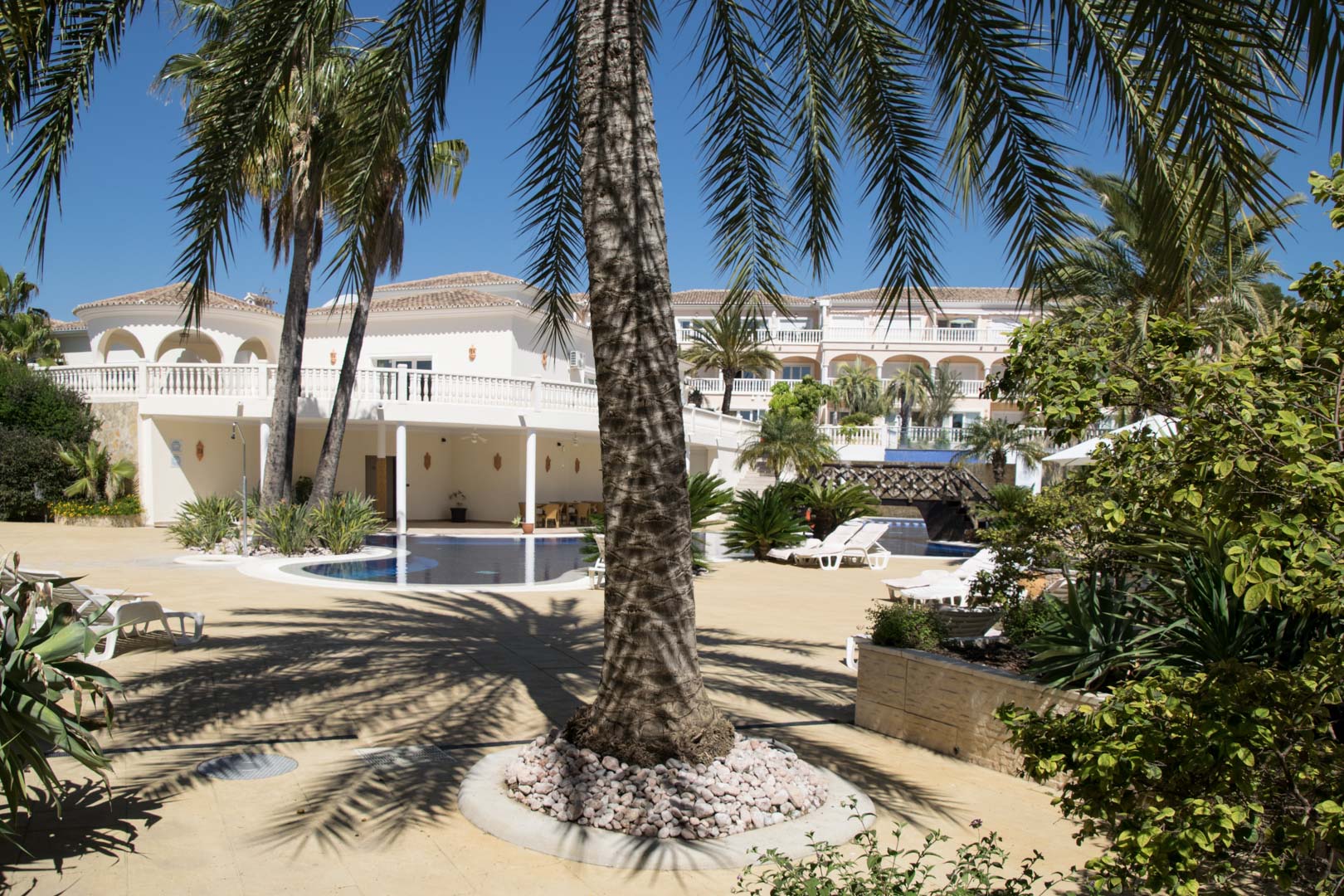 Buitenruimte met lagunezwembad, omgeven door palmbomen en mediterrane tuinen. De architectuur kenmerkt zich door witte gevels, balustrades en zonnige terrassen.