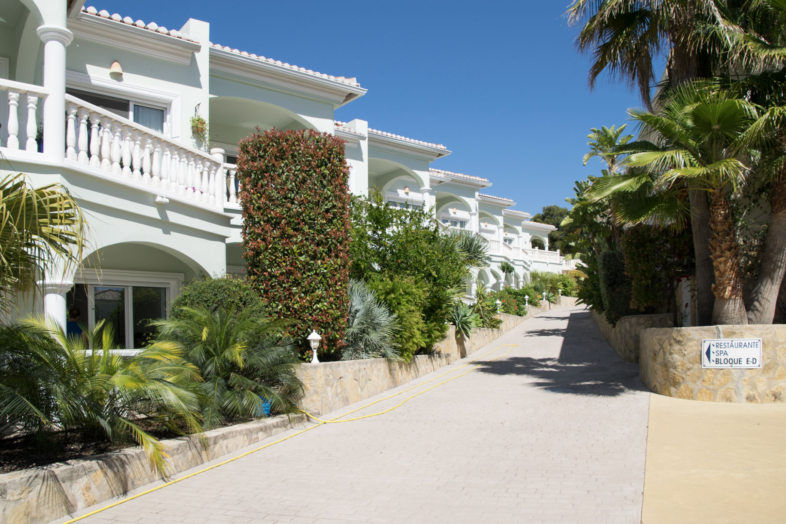 Exterieur van het mediterrane complex met witte balustrades, gewelfde terrassen en weelderige beplanting langs een verzorgd geplaveid wandelpad.