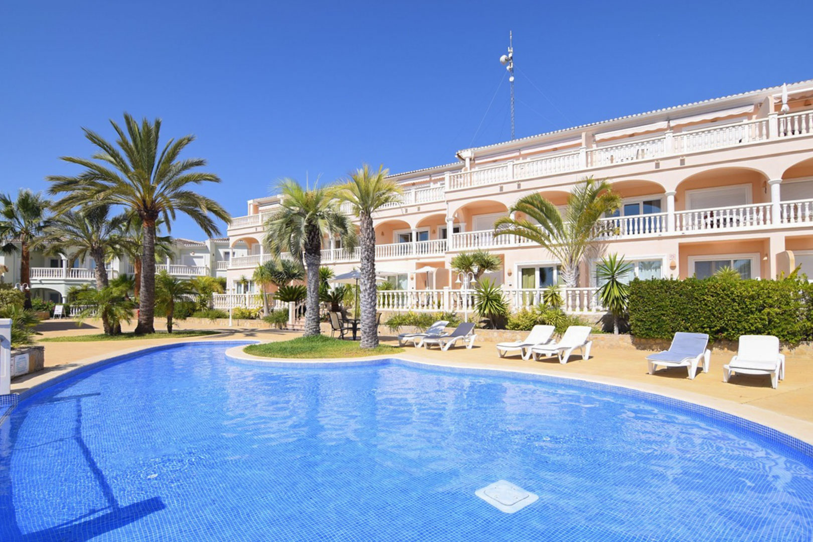 Gemeenschappelijk lagune-zwembad met blauwe tegels, omgeven door palmbomen en ligbedden voor een mediterraan gebouw met boogvormige terrassen en witte balustrades.