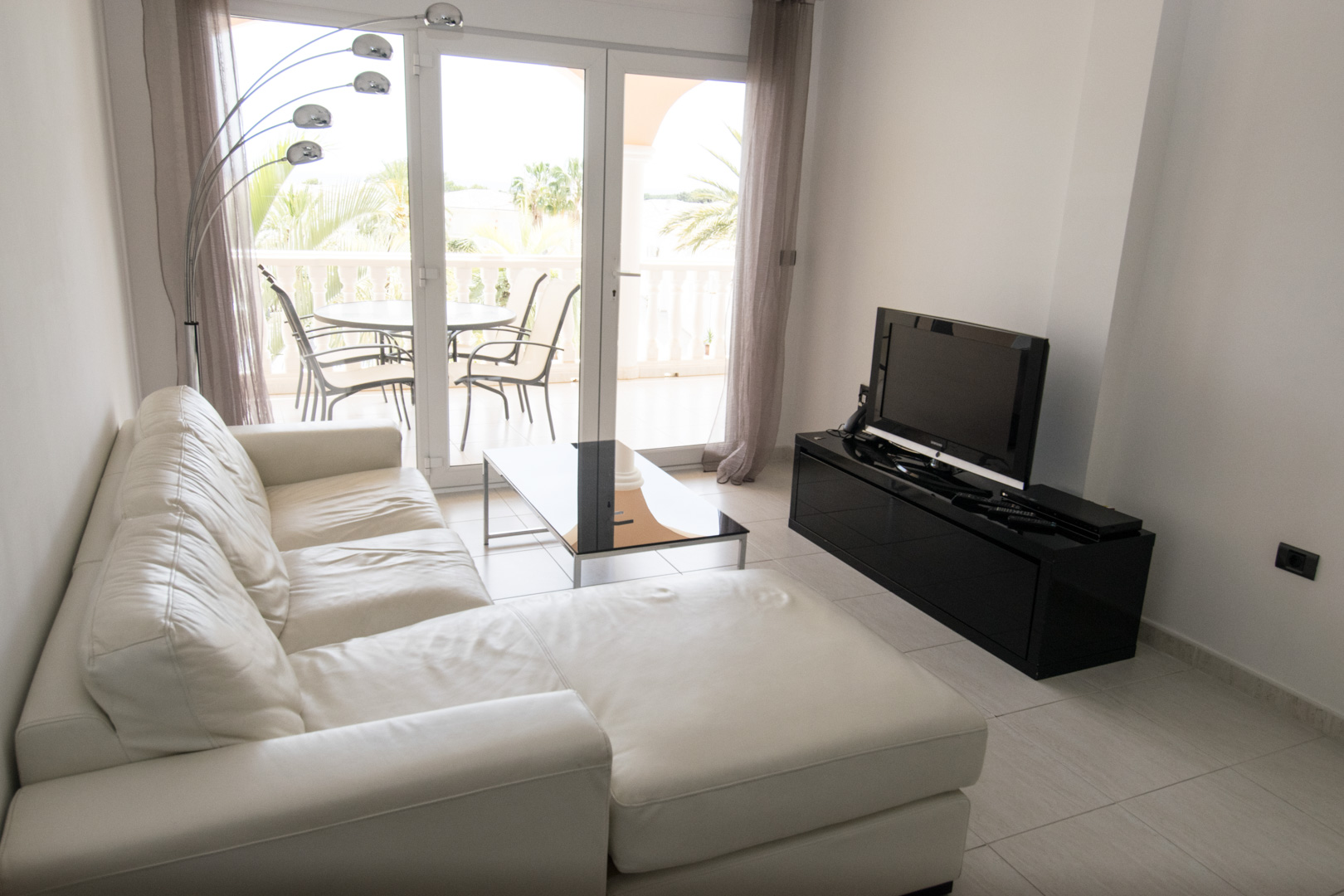 Lichte woonkamer met een wit lederen chaise longue, zwart hoogglans tv-meubel en glazen schuifpui naar een terras met uitzicht op palmbomen.