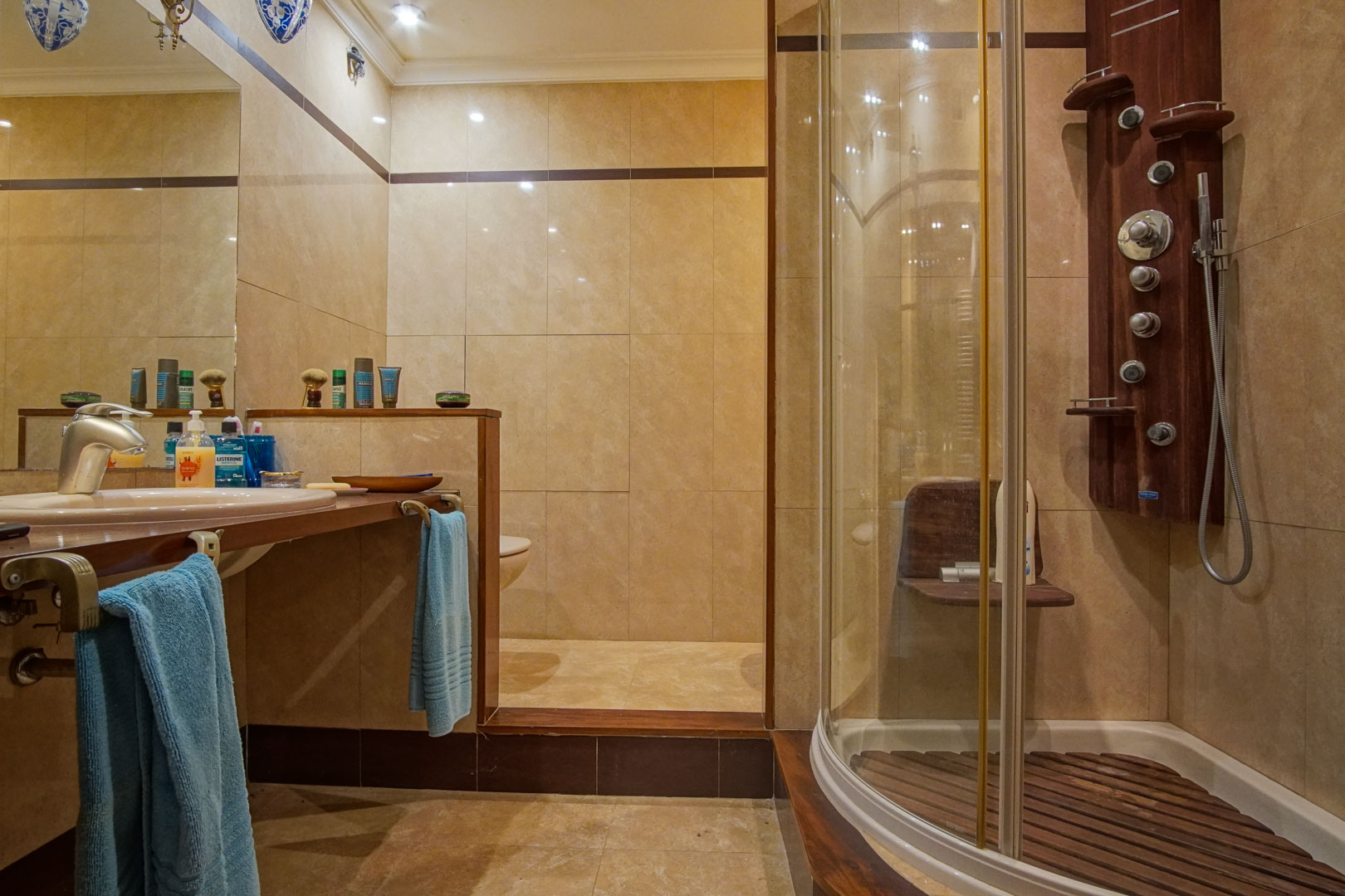 Badkamer met beige marmeren tegels, een houten wastafelblad en een halfronde douchecabine met hydromassagekolom en houten vlonder.