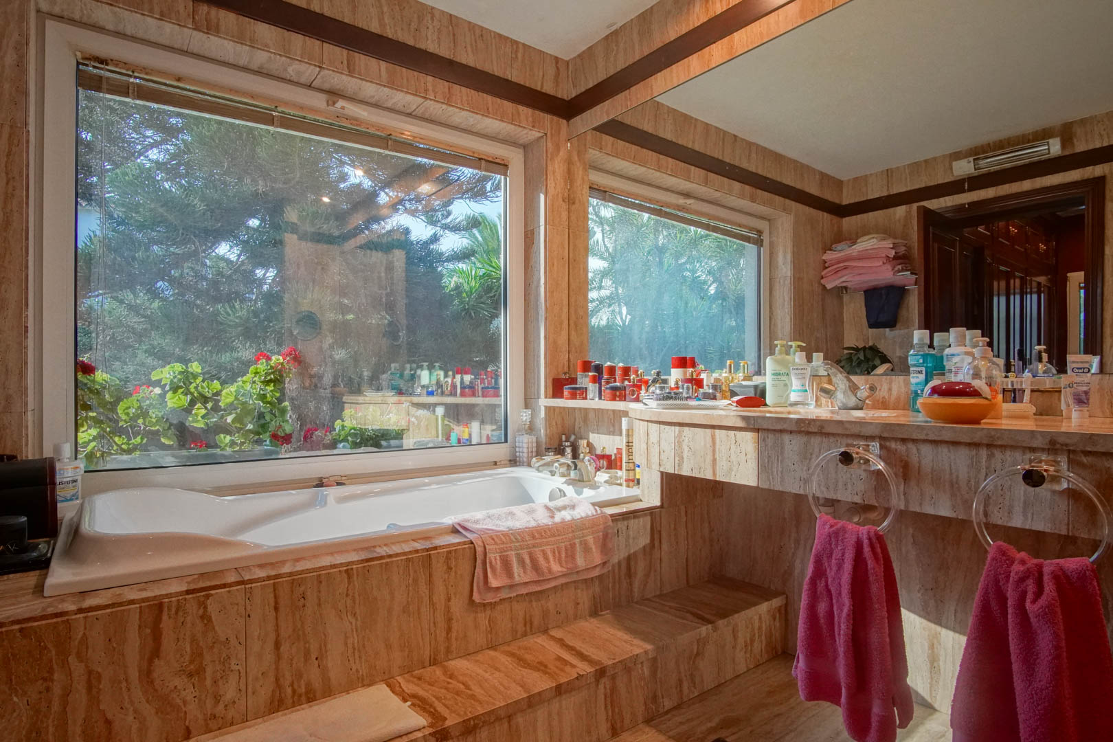 Badkamer met travertijn marmeren bekleding, een ingebouwd bad bij een groot raam met uitzicht op de tuin en een geïntegreerd wastafelmeubel met grote spiegel.