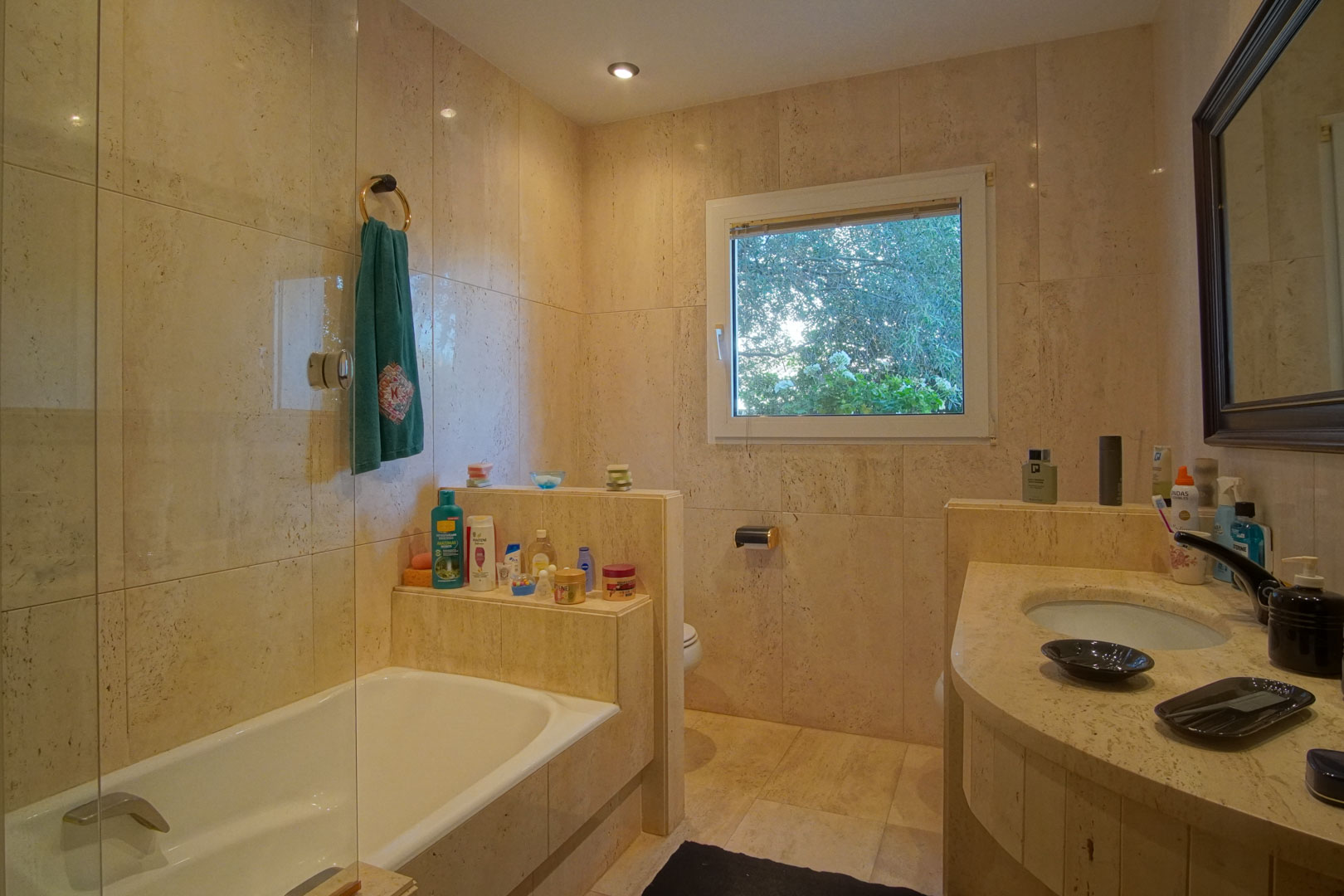 Badkamer volledig bekleed met crème marmer, voorzien van een ligbad met glazen wand en een natuurstenen wastafelblad. Het raam biedt natuurlijk licht en uitzicht.