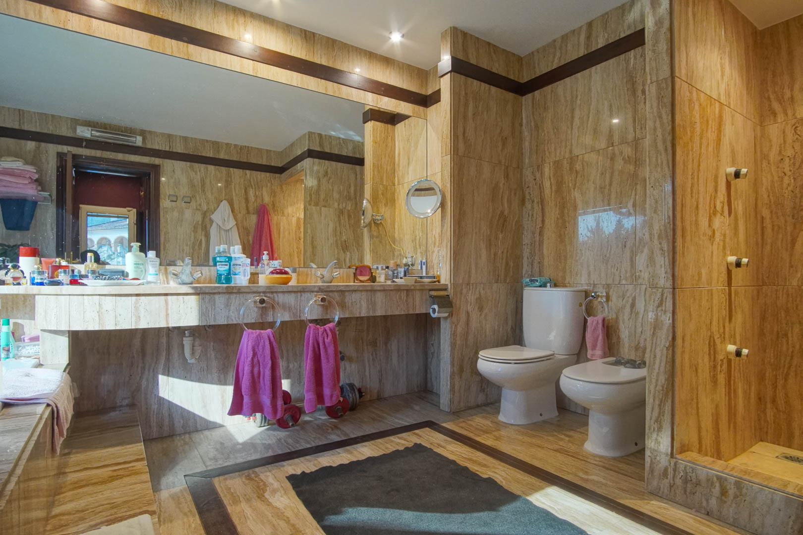 Badkamer volledig bekleed met travertijn marmer, voorzien van een geïntegreerd wastafelmeubel, grote spiegel en subtiele inbouwverlichting.