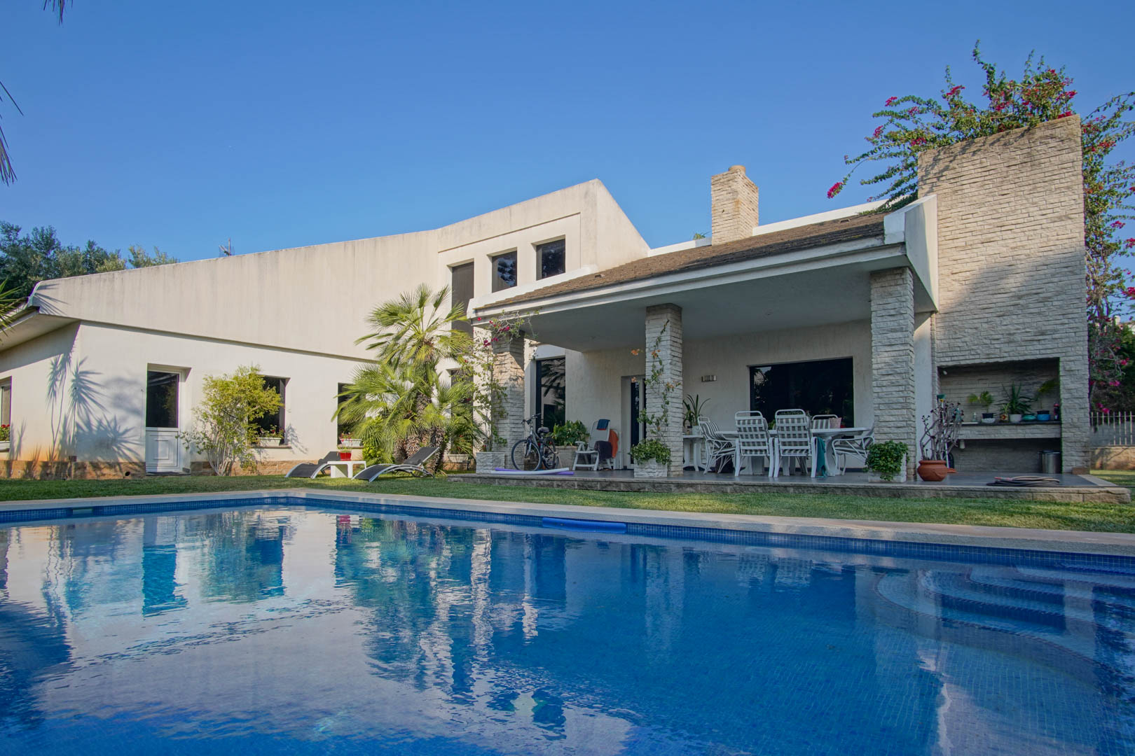 Buitenaanzicht van het privézwembad en overdekt terras met stenen zuilen, buiten-eethoek en geïntegreerde barbecue in een mediterrane tuin.